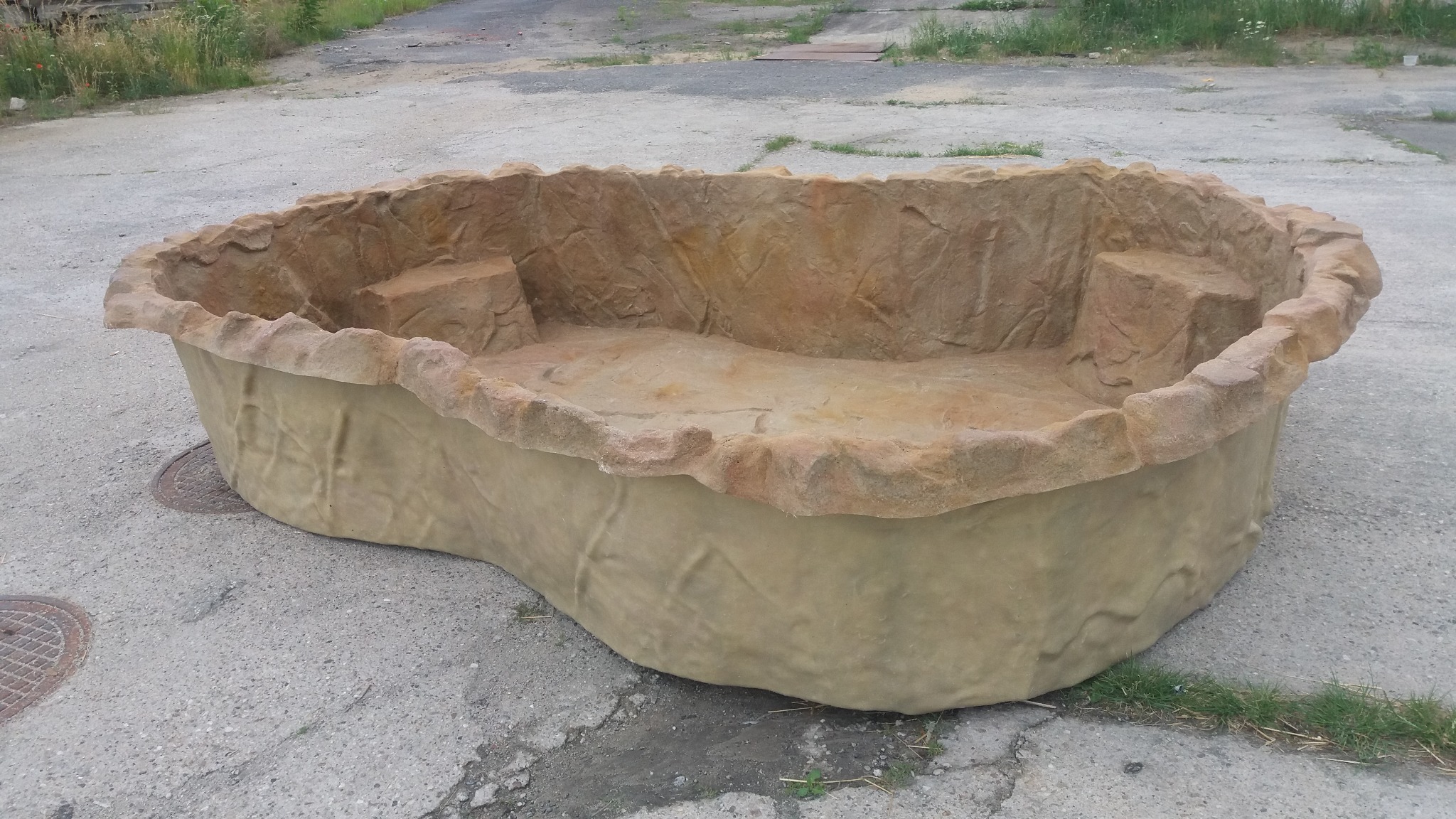 Gotowy, beżowy zbiornik imitujący kamień, przeznaczony do budowy oczka wodnego. Widoczne dwa stopnie wewnątrz. Tło stanowi betonowa nawierzchnia.