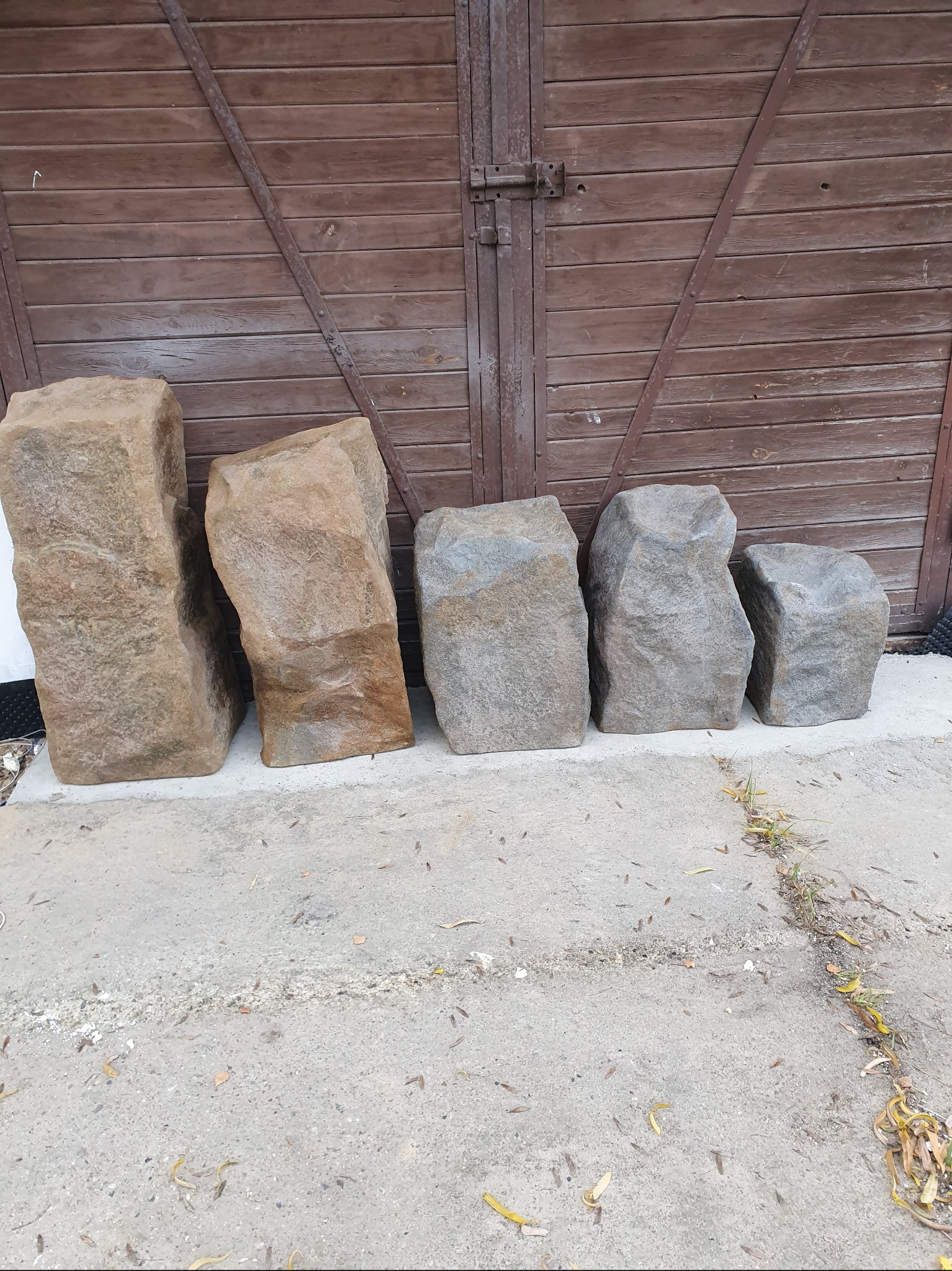 Różnej wielkości bloki skalne w odcieniach beżu i szarości, ustawione rzędem na betonowej powierzchni przed brązowymi, drewnianymi drzwiami.