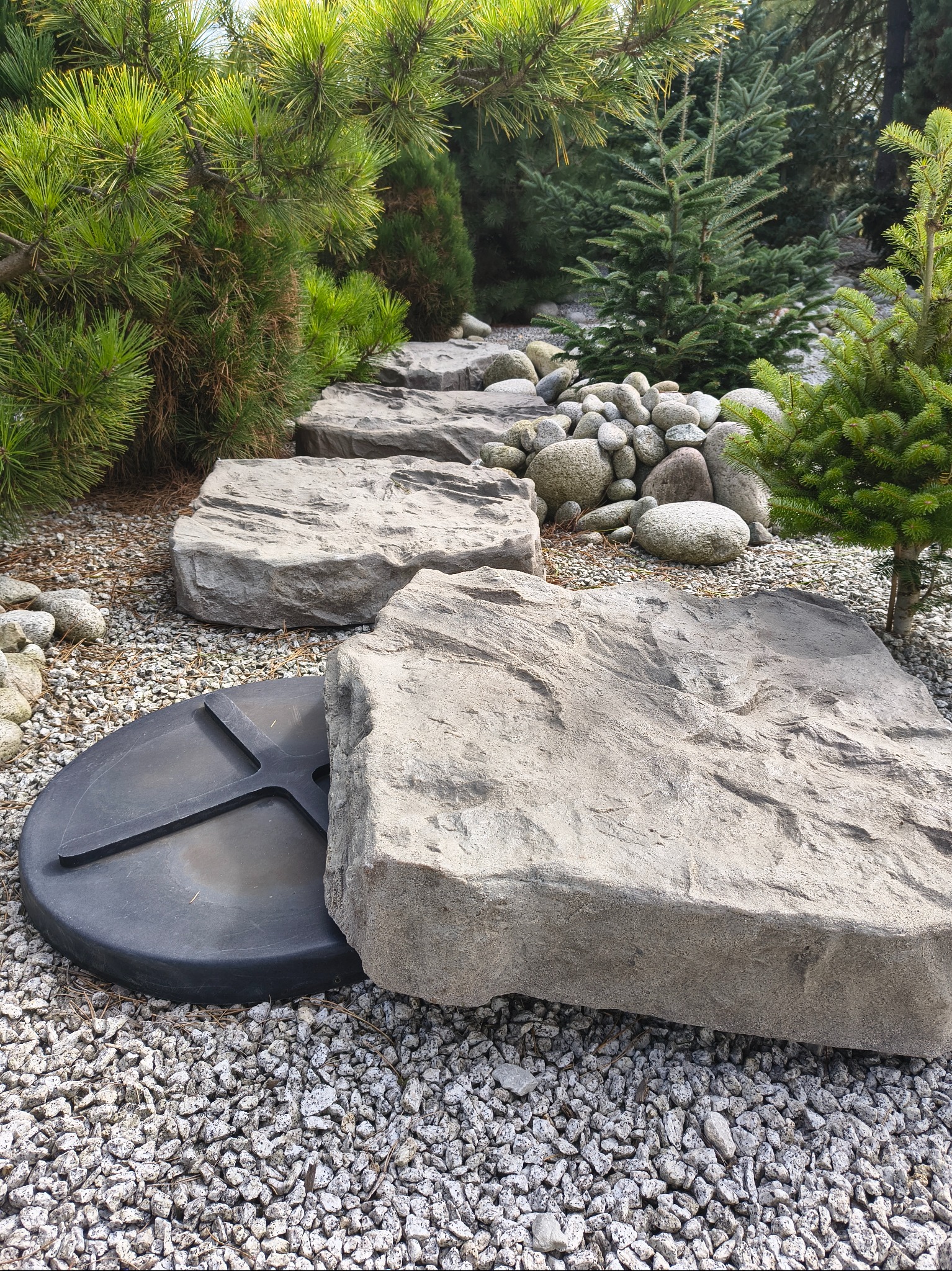 Kamienne płyty ścieżki ogrodowej, częściowo zakrywają czarną pokrywę studzienki, otoczone żwirem i zielenią iglaków.