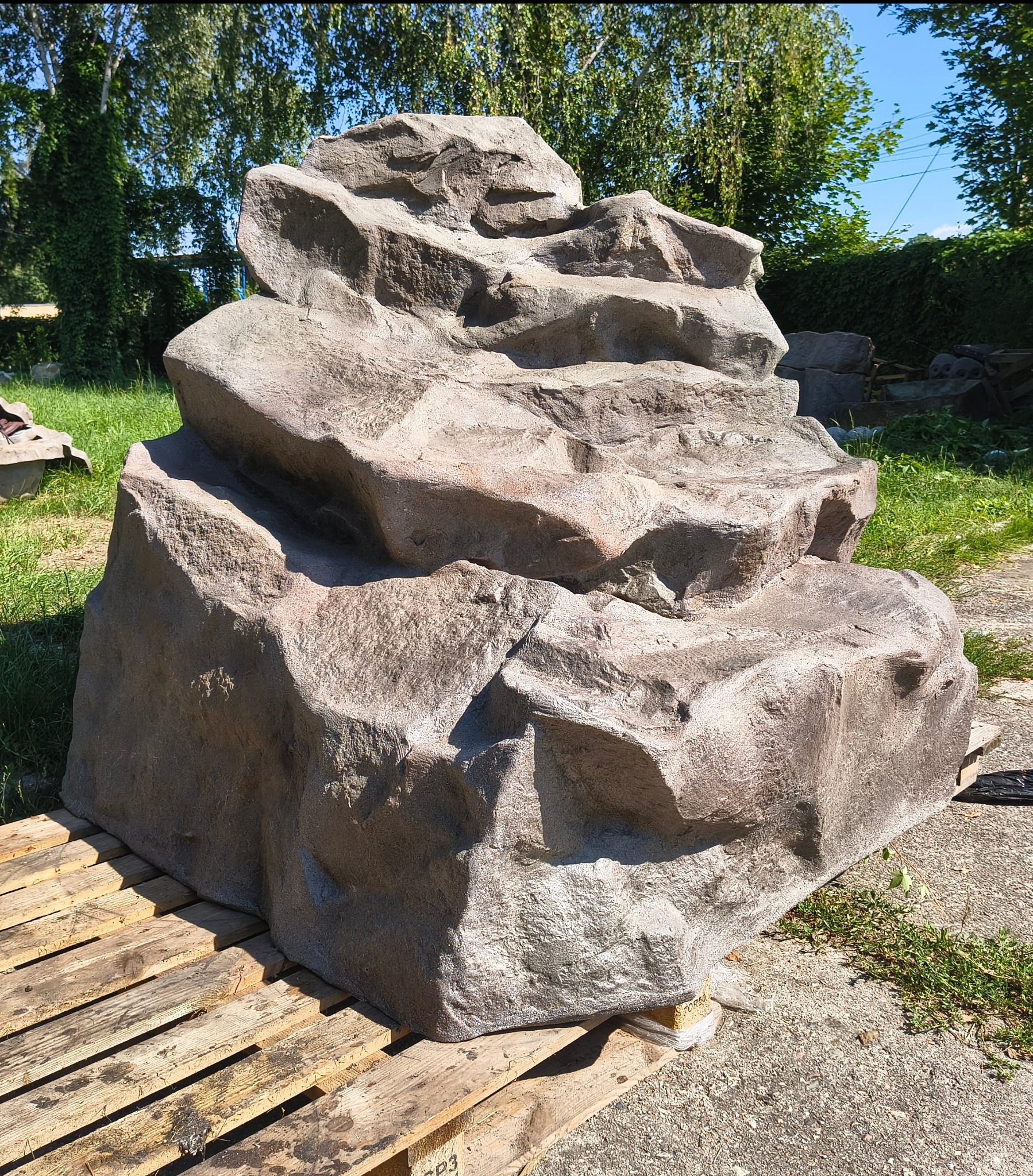 Dekoracyjny, sztuczny wodospad z kamienia na palecie, imitujący naturalną skałę, z zielenią w tle. Element aranżacji przestrzeni zewnętrznej.