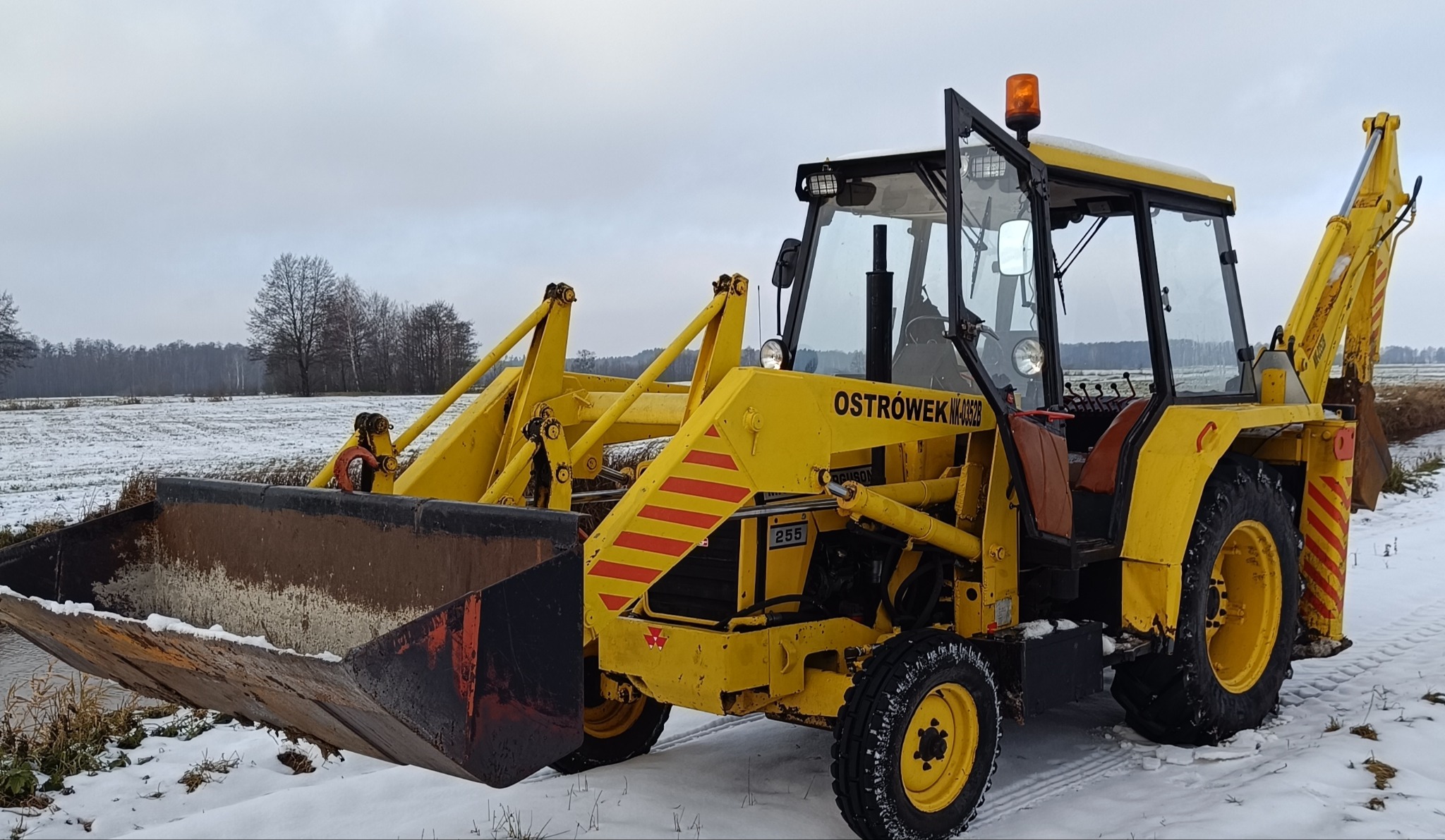 Żółta koparko-ładowarka Ostrowek NK-0352B na zasypanym śniegiem polu. Widoczne ramię koparki, łyżka i kabina. Na maszynie czerwone pasy i oznaczenie 255.