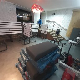 Michał Sikora - Wnętrze lokalu w trakcie wyposażania: ustawione rzędy ławek z oparciami z białych listew, zapakowane siedziska, schody w tle, dekoracyjna lampa.