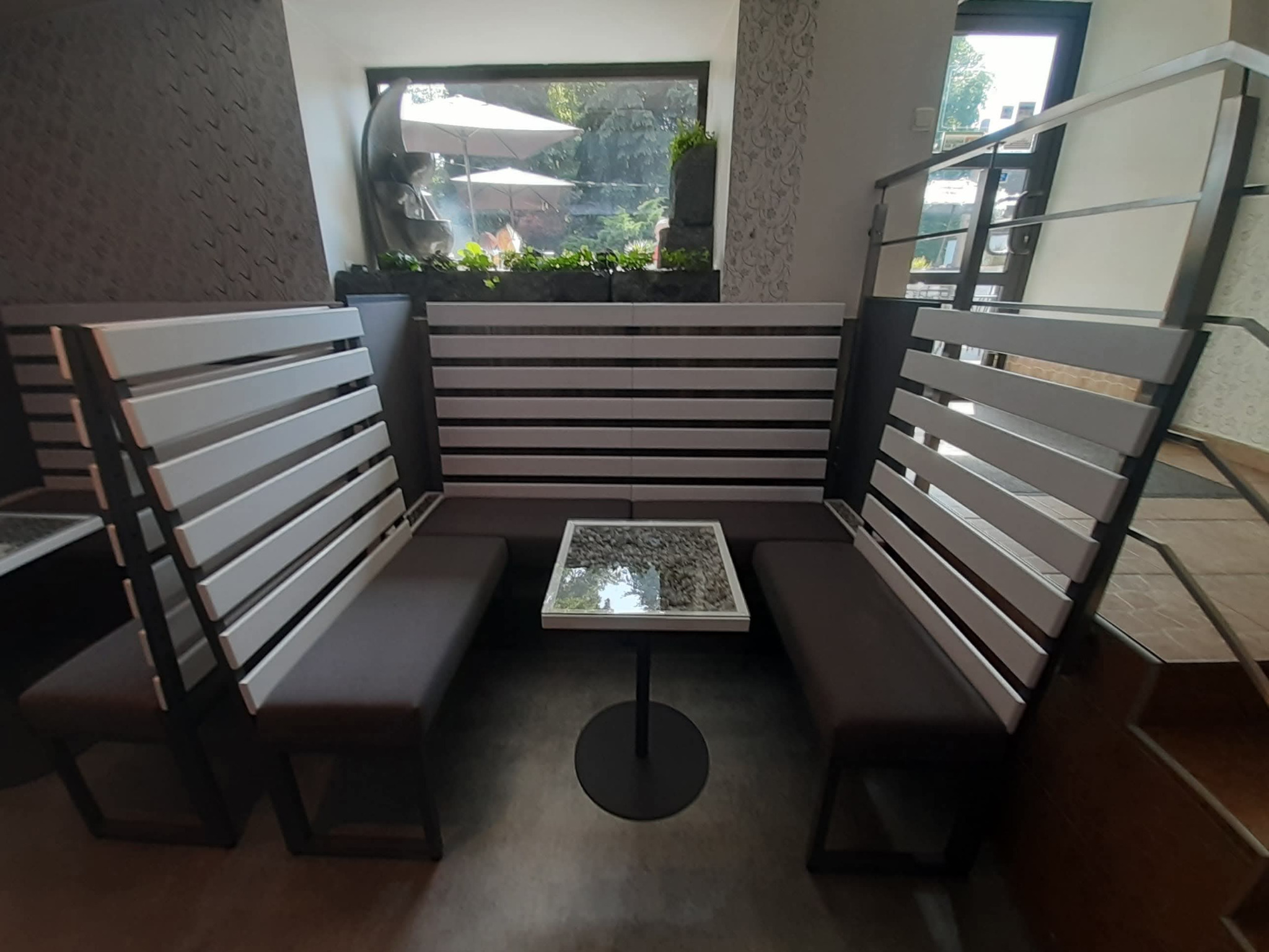 Nowoczesna zabudowa siedzisk z drewnianymi panelami i stolikiem w lokalu gastronomicznym w Kętach. Widok na okno z zielenią i parasolami.