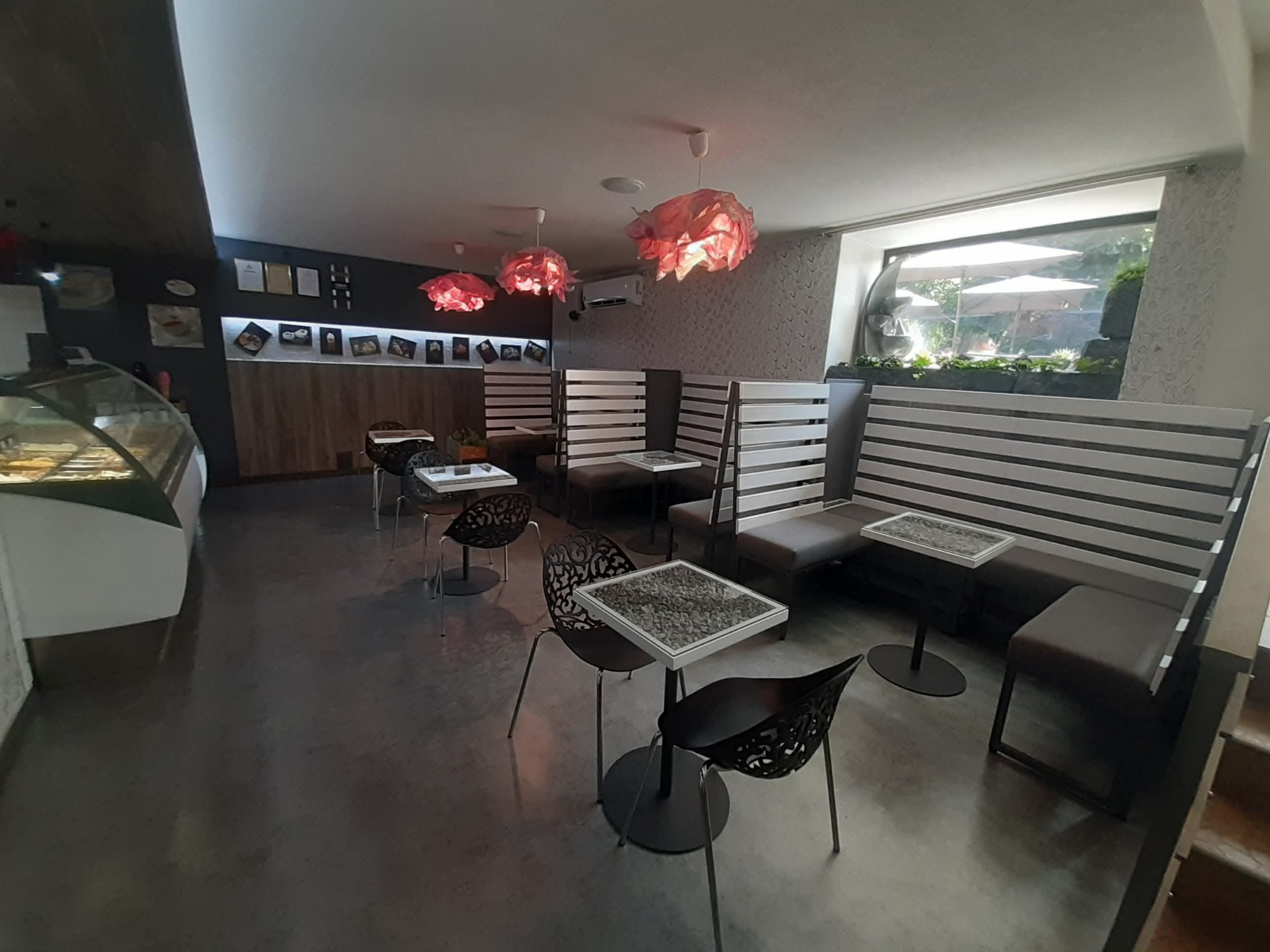 Wnętrze kawiarni z szarymi podłogami, designerskimi krzesłami i stolikami z kamiennymi blatami. W tle loże z drewnianymi panelami i widok na ogród przez okno.