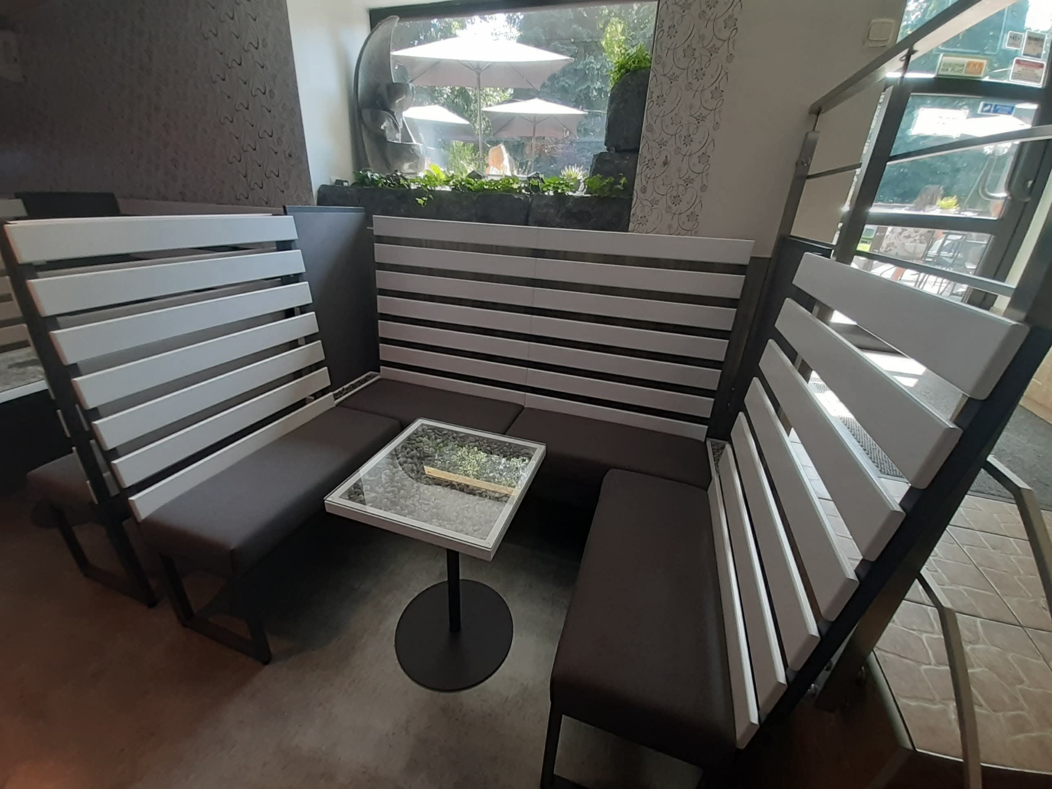 Nowoczesna strefa relaksu w lokalu gastronomicznym: grafitowe siedziska, białe lamele, szklany stolik na tle okna z widokiem na zieleń i fontannę.