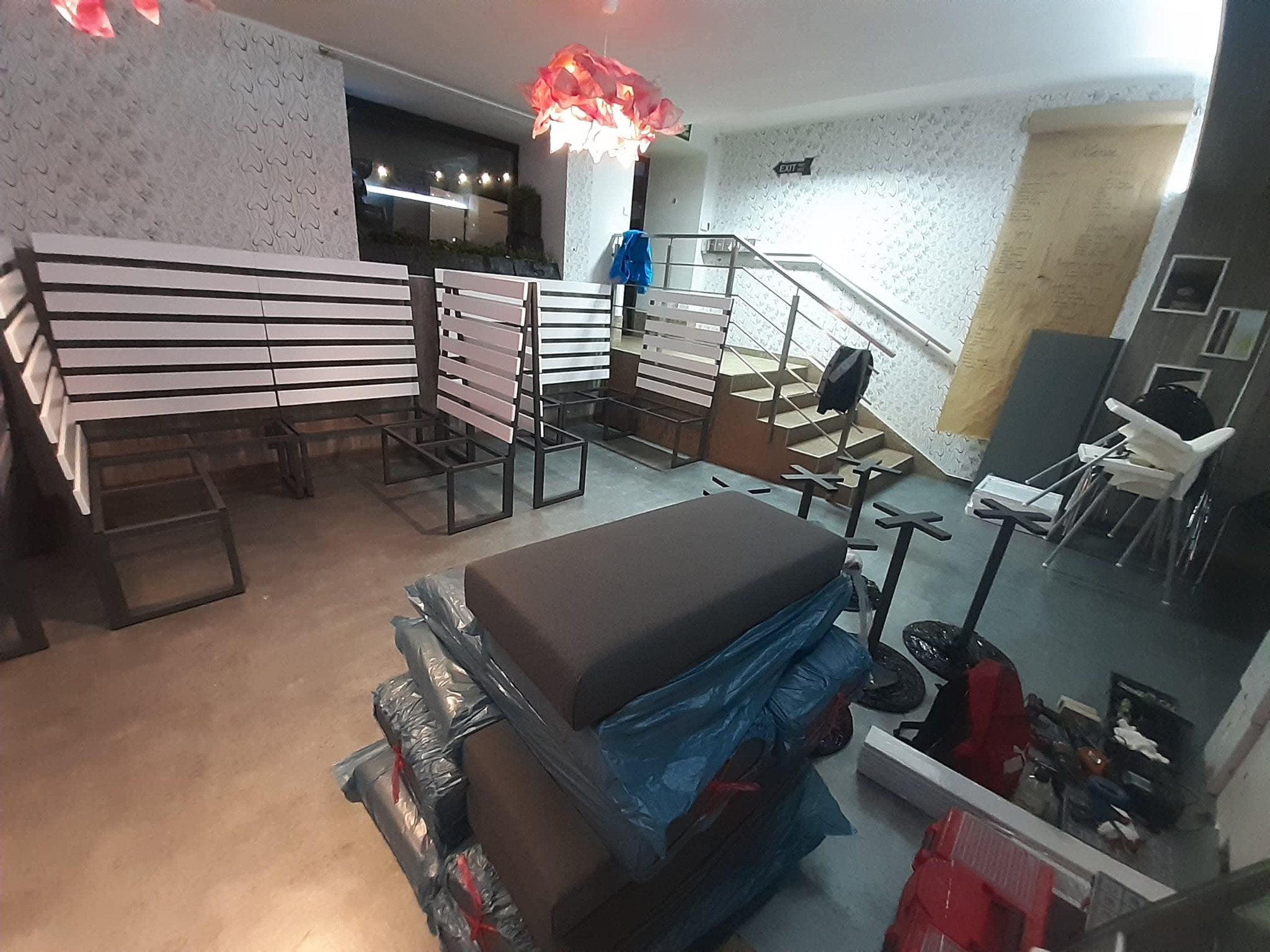 Wnętrze lokalu w trakcie wyposażania: ustawione rzędy ławek z oparciami z białych listew, zapakowane siedziska, schody w tle, dekoracyjna lampa.