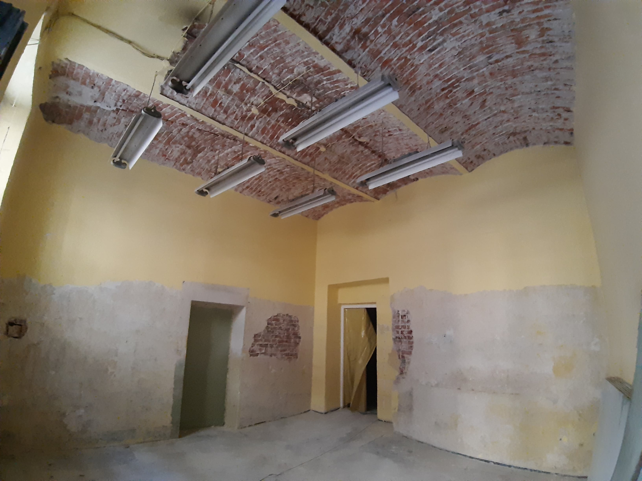 Wnętrze w trakcie remontu: odsłonięta cegła na sklepieniu łukowym, surowe ściany z fragmentami starego tynku, widoczne instalacje oświetleniowe i otwory drzwiowe.