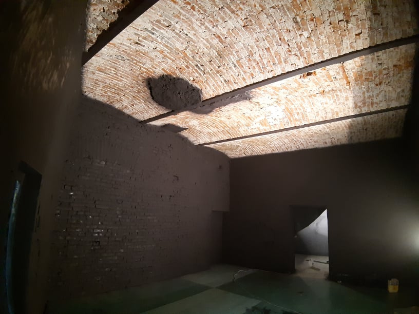 Wnętrze w trakcie prac wykończeniowych: otynkowane ściany z cegły, sklepienie łukowe z cegły, widoczne metalowe belki stropowe i fragment podłogi z narzędziami.
