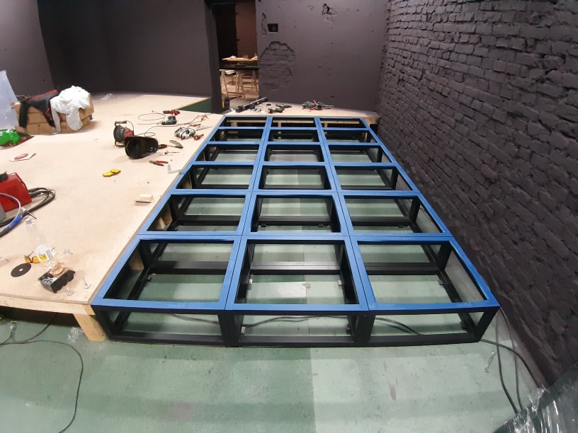 Metalowa konstrukcja pod podest sceniczny z niebieskimi akcentami, w surowym wnętrzu z ceglaną ścianą. Widoczne narzędzia i elementy montażowe.