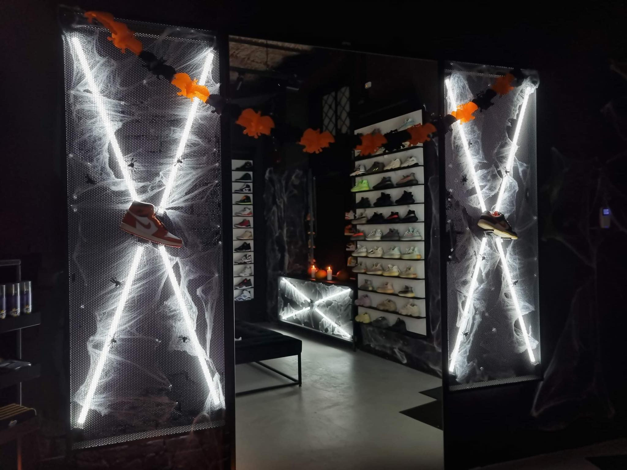 Wnętrze sklepu obuwniczego z dekoracjami Halloween: pajęczyny, dynie i nietoperze. Podświetlane panele z butami sportowymi w tle. Mroczny klimat.