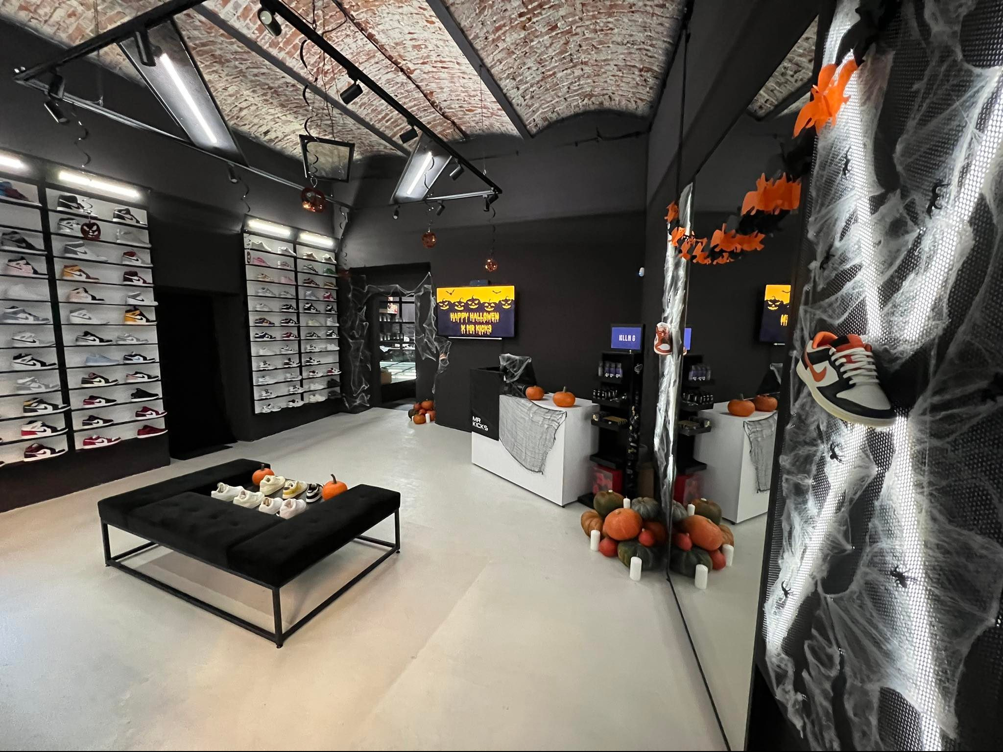 Wnętrze sklepu z butami sportowymi, dekoracje Halloween: dynie, pajęczyny, nietoperze. Ekspozycja obuwia na półkach i na czarnej ławie. Ceglany sufit.