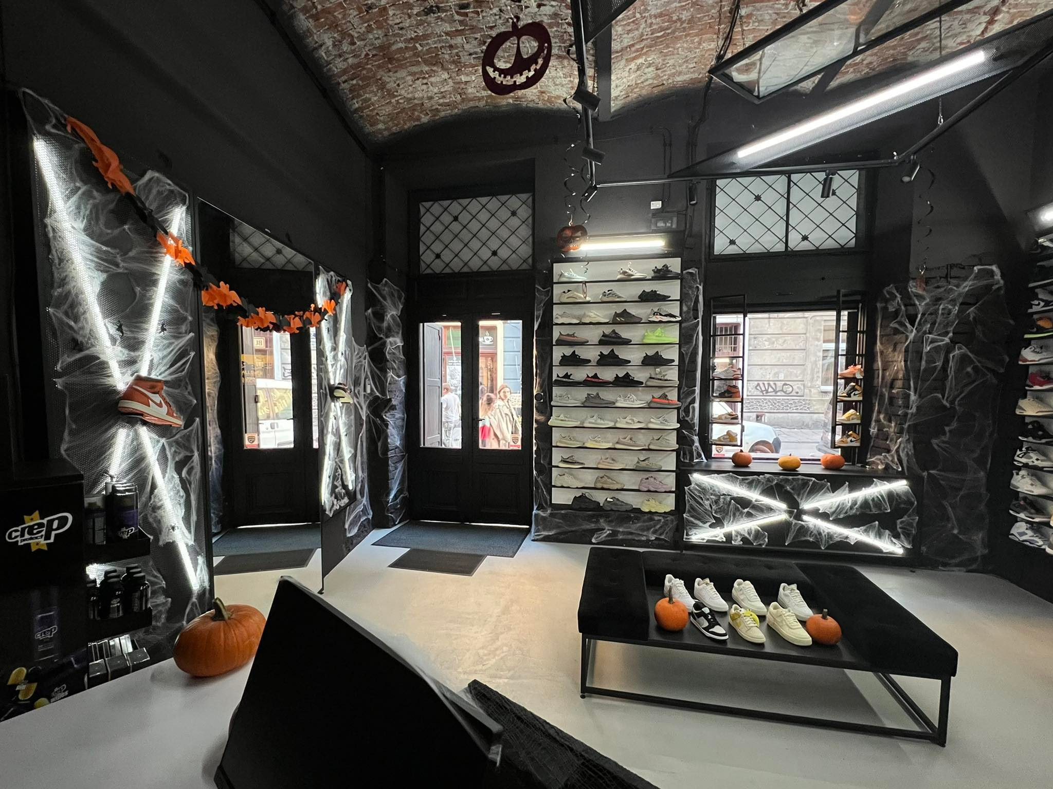 Ciemne wnętrze sklepu obuwniczego z dekoracjami Halloween: pajęczyny, dynie, girlandy. Ekspozycja butów na półkach i stoliku. Ceglany sufit i czarne ściany.