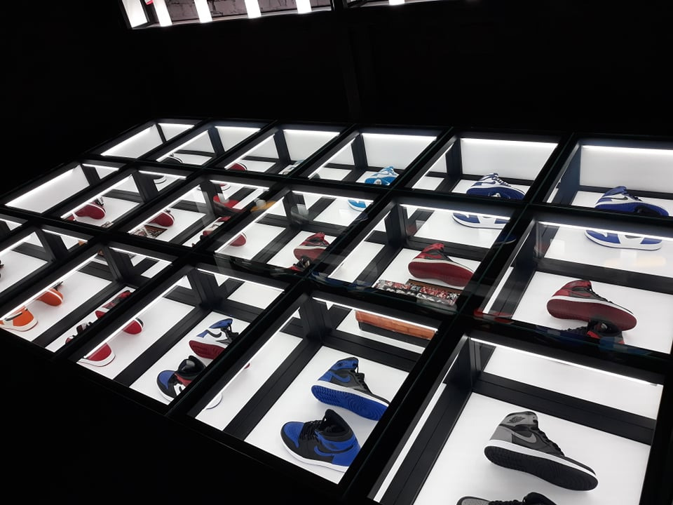 Podświetlana ekspozycja butów sportowych w czarnych, geometrycznych gablotach. Różne modele i kolory, ułożone w regularnych odstępach, tworzą nowoczesny design.
