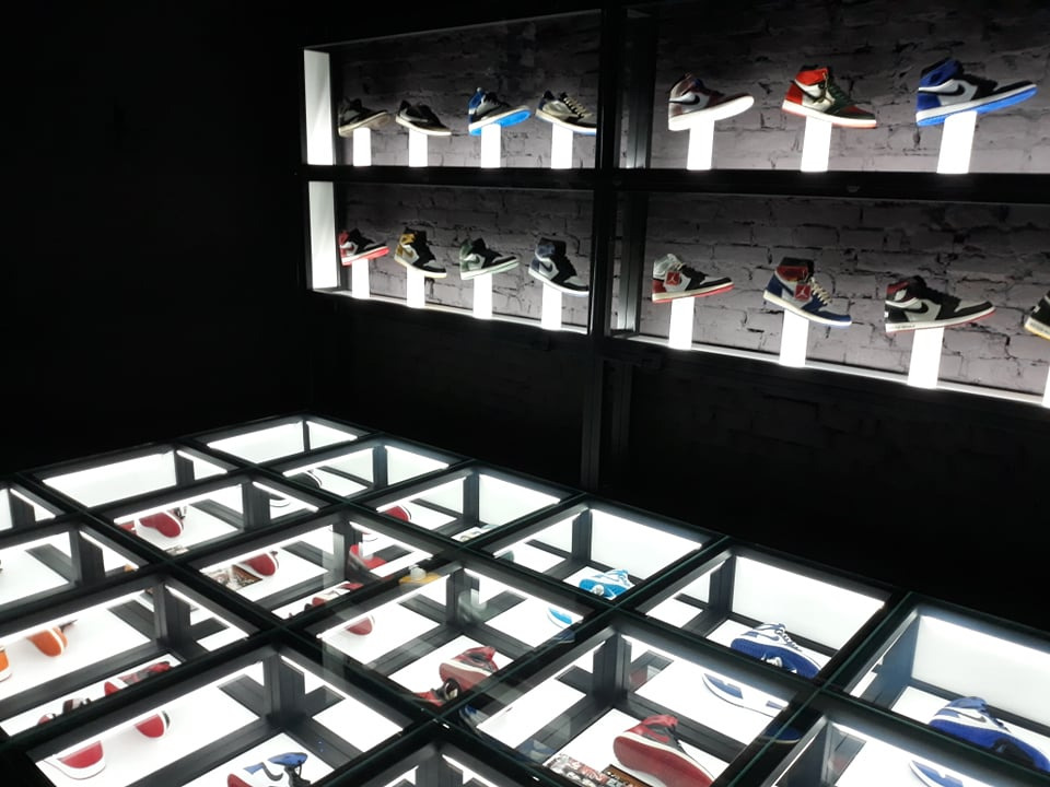 Oświetlona ekspozycja butów sportowych w sklepie: półki ścienne z podświetleniem i przeszklona podłoga z butami. Ciemne tło, ceglana ściana.