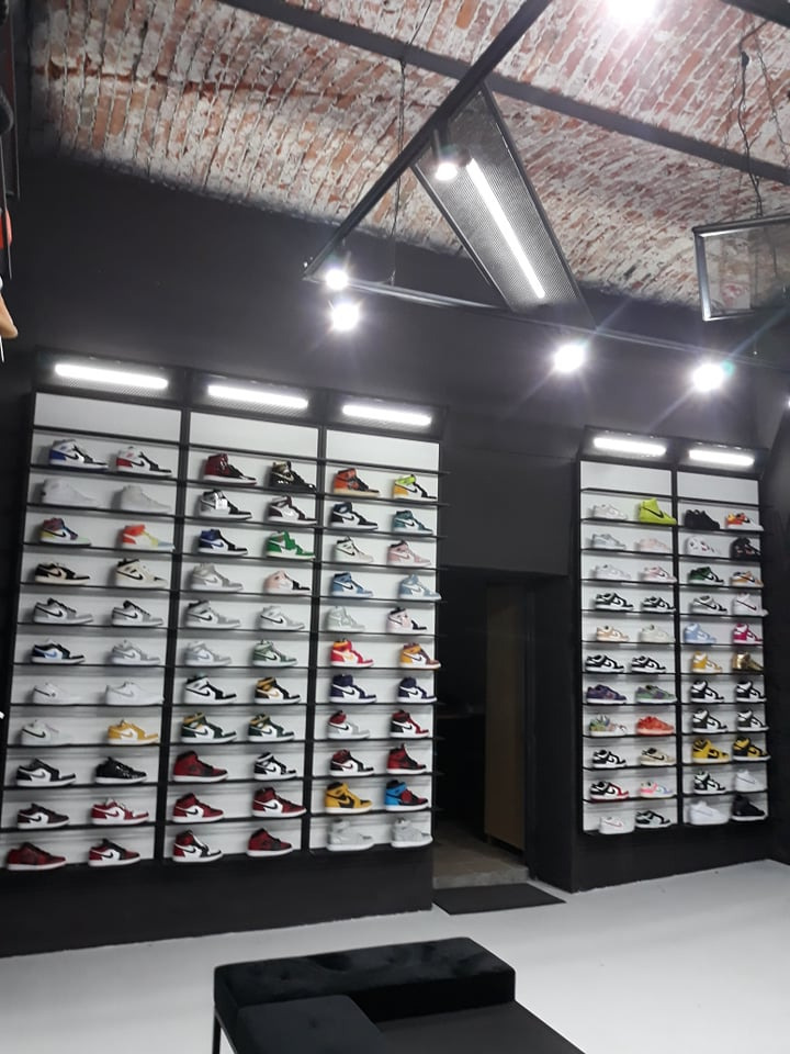 Wnętrze sklepu obuwniczego z czarnymi ścianami i ceglanym sufitem. Na ścianach półki z kolekcją kolorowych butów sportowych.