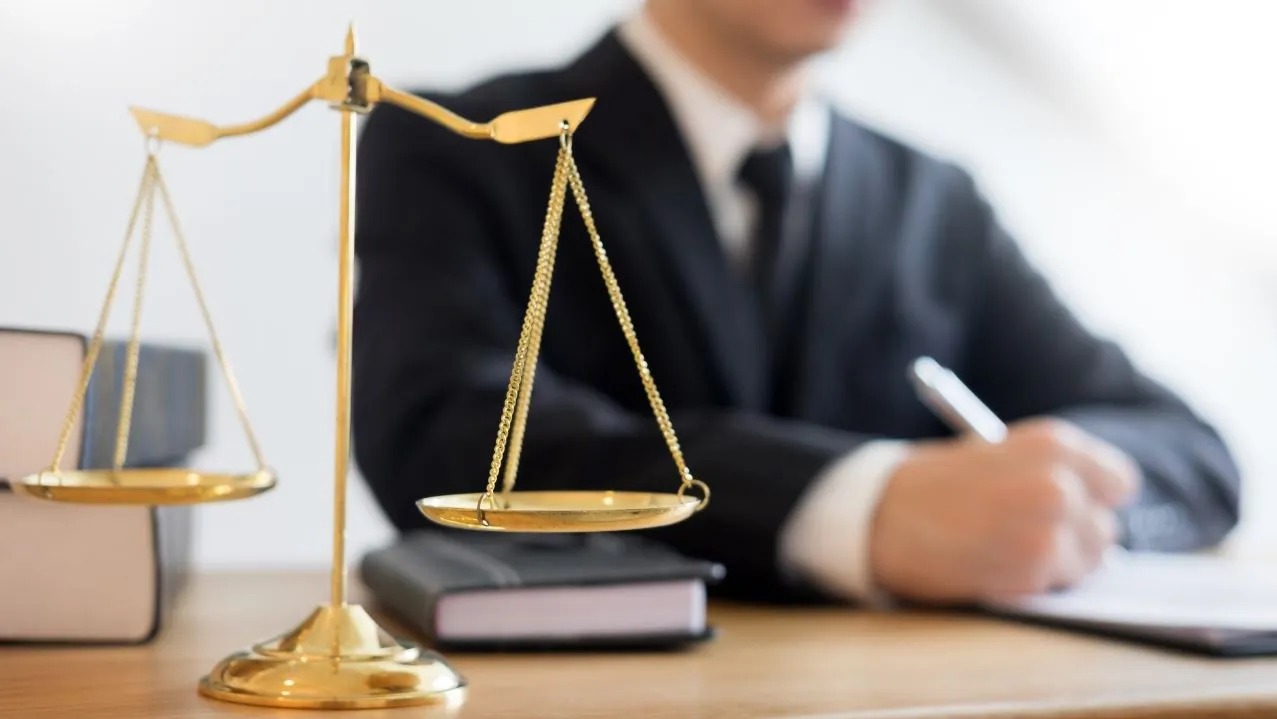 Złote wagi Temidy na biurku, w tle prawnik w garniturze sporządzający notatki. Symbol sprawiedliwości i prawa w kancelarii.