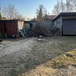 GreenCut - Sterta gałęzi i pni po wycince drzew na posesji, widoczne garaże i ogrodzenie. Słoneczny dzień.