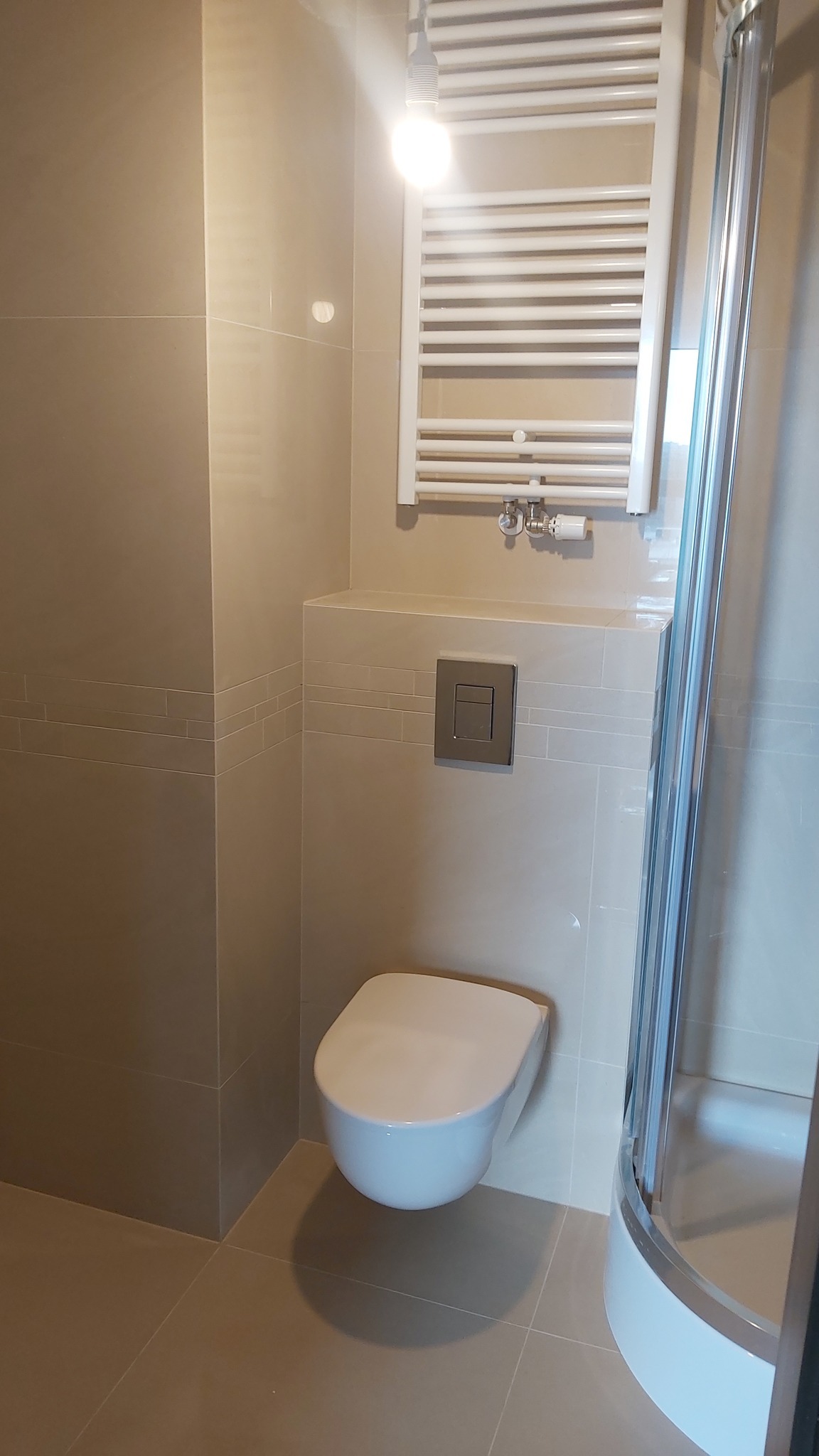 Beżowa łazienka z podwieszaną toaletą, grzejnikiem drabinkowym i kabiną prysznicową. Minimalistyczny design, płytki w odcieniach beżu, nowoczesna armatura. Oświetlenie górne.