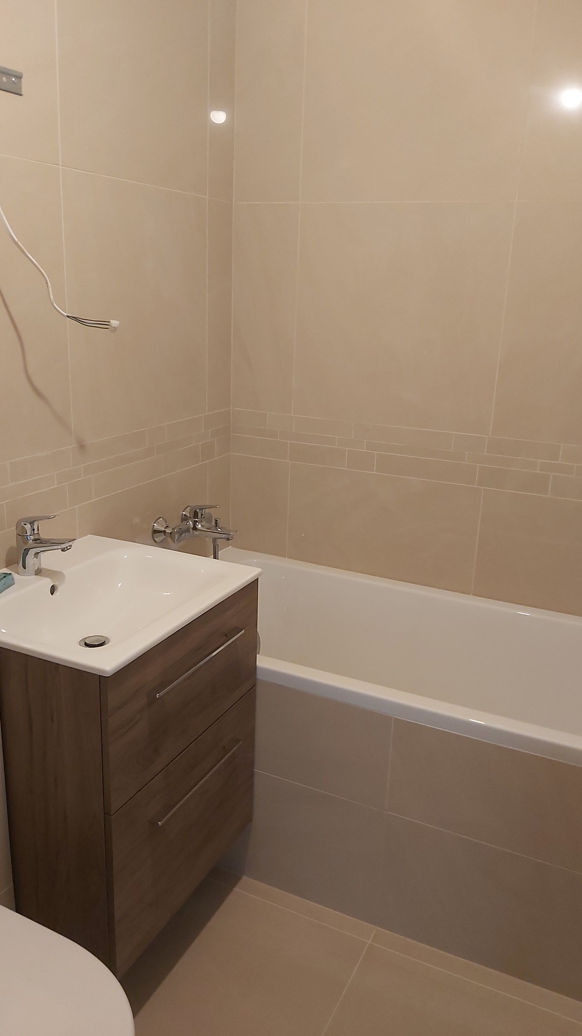 Jasna łazienka z beżowymi płytkami, umywalką z szafką w kolorze drewna i wanną. Minimalistyczny design, chromowana armatura, ciepłe oświetlenie.