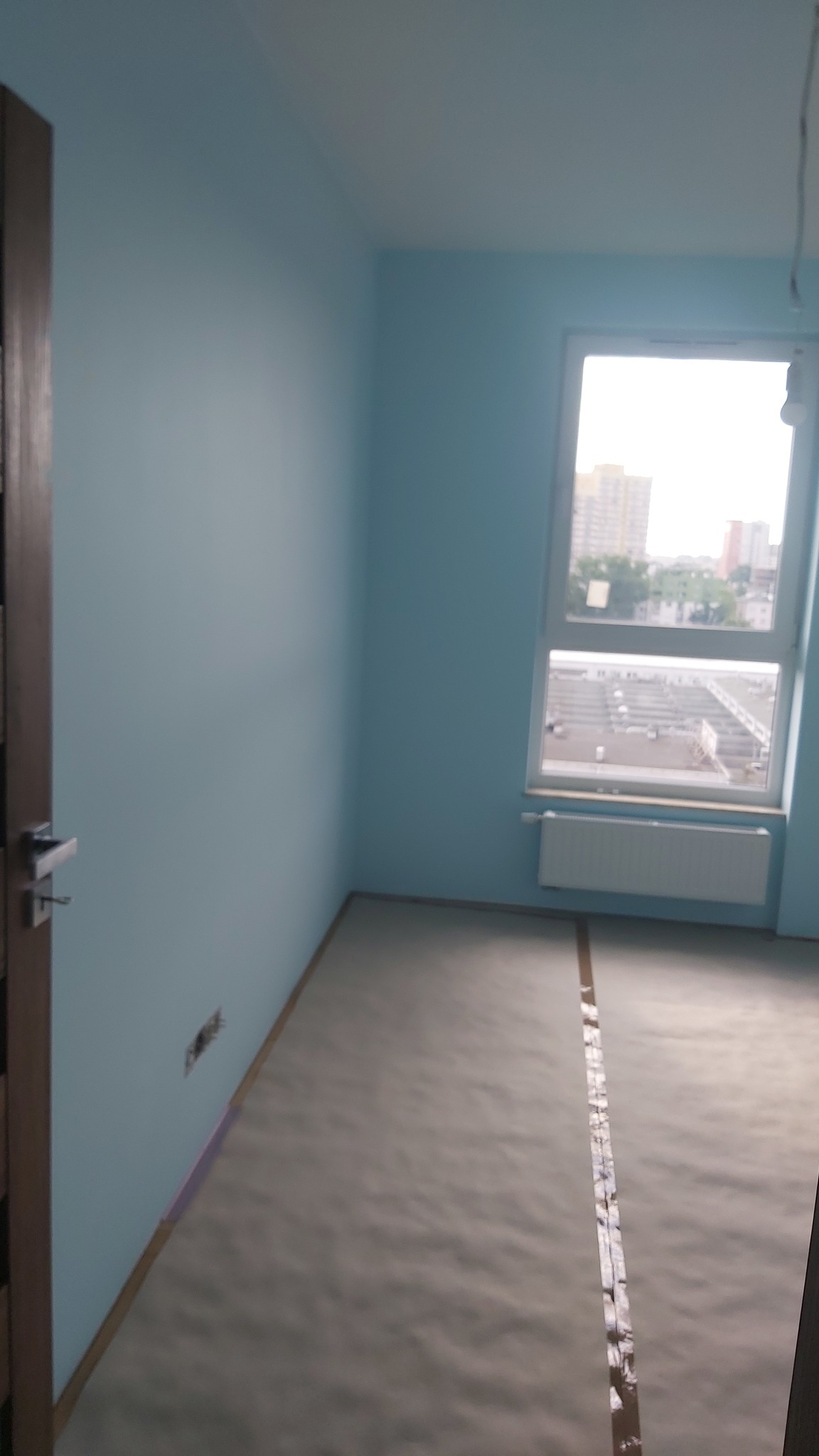 Pomalowany na błękitno pokój z oknem i zabezpieczoną podłogą. Widoczne ślady po gniazdku elektrycznym. Przygotowanie do dalszych prac wykończeniowych.