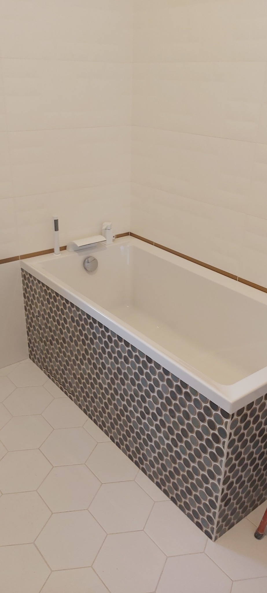 Biała wanna z mozaiką w łazience z heksagonalną podłogą i kremowymi płytkami ściennymi z delikatną fakturą. Widoczny kran i słuchawka prysznicowa.