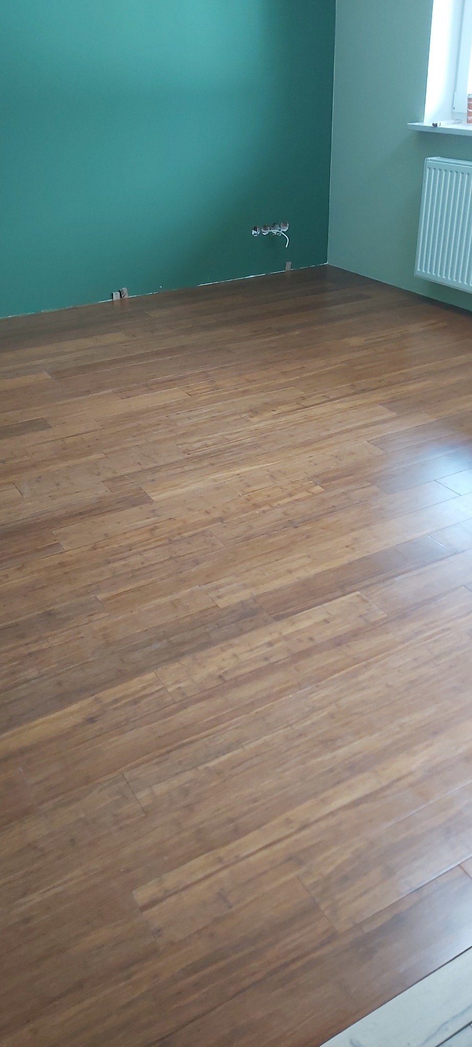 Ułożony parkiet w pokoju z zielonymi ścianami i grzejnikiem. Widoczne gniazdka elektryczne. Podłoga z paneli w odcieniach brązu. Jasne wnętrze po remoncie.