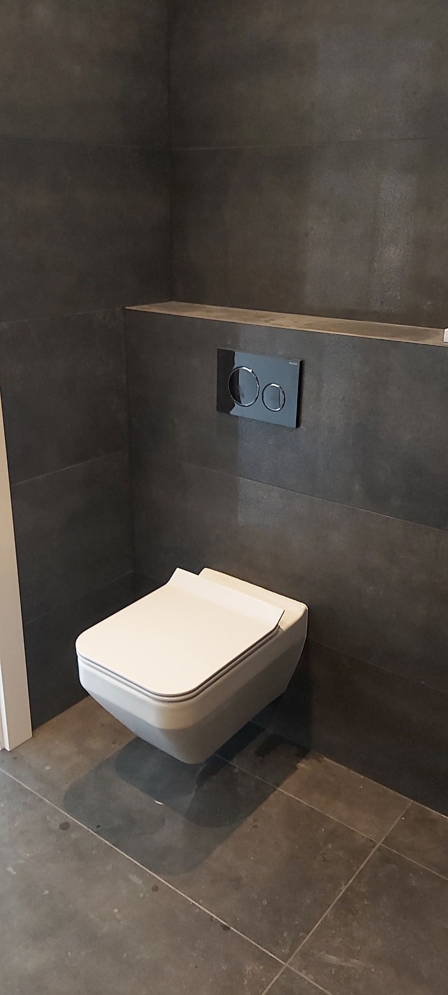 Nowoczesna toaleta podwieszana z szarą płytką na ścianach i podłodze. Minimalistyczny design, czarny przycisk spłukujący. Wykończona łazienka w ciemnych tonacjach.