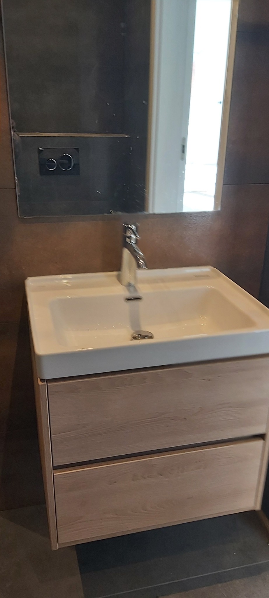 Nowoczesna łazienka: biała umywalka z drewnianą szafką, chromowana bateria, lustro z widocznym fragmentem toalety podtynkowej i ciemne płytki na ścianie.