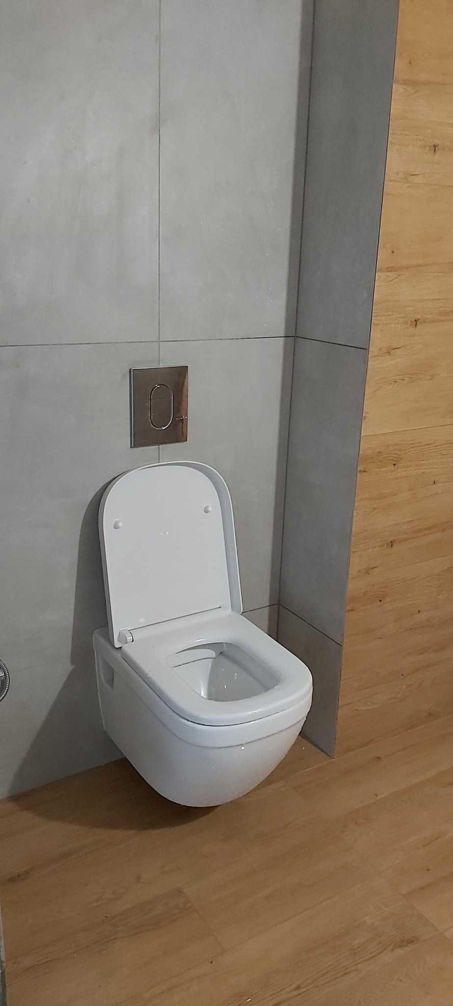 Nowoczesna toaleta podwieszana z chromowanym przyciskiem spłukującym na szarej, betonowej ścianie, uzupełnona drewnopodobną podłogą i ścianą.