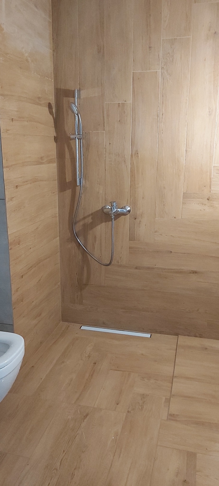 Wykończona łazienka w Warszawie z drewnopodobnymi płytkami, chromowaną armaturą i odpływem liniowym. Minimalistyczny design, ciepłe oświetlenie.