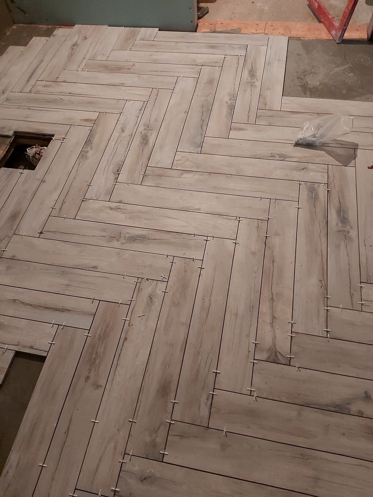 Ułożone płytki podłogowe w jodełkę, imitujące drewno, z krzyżykami dystansowymi. Widoczny otwór w podłodze i worek z krzyżykami.