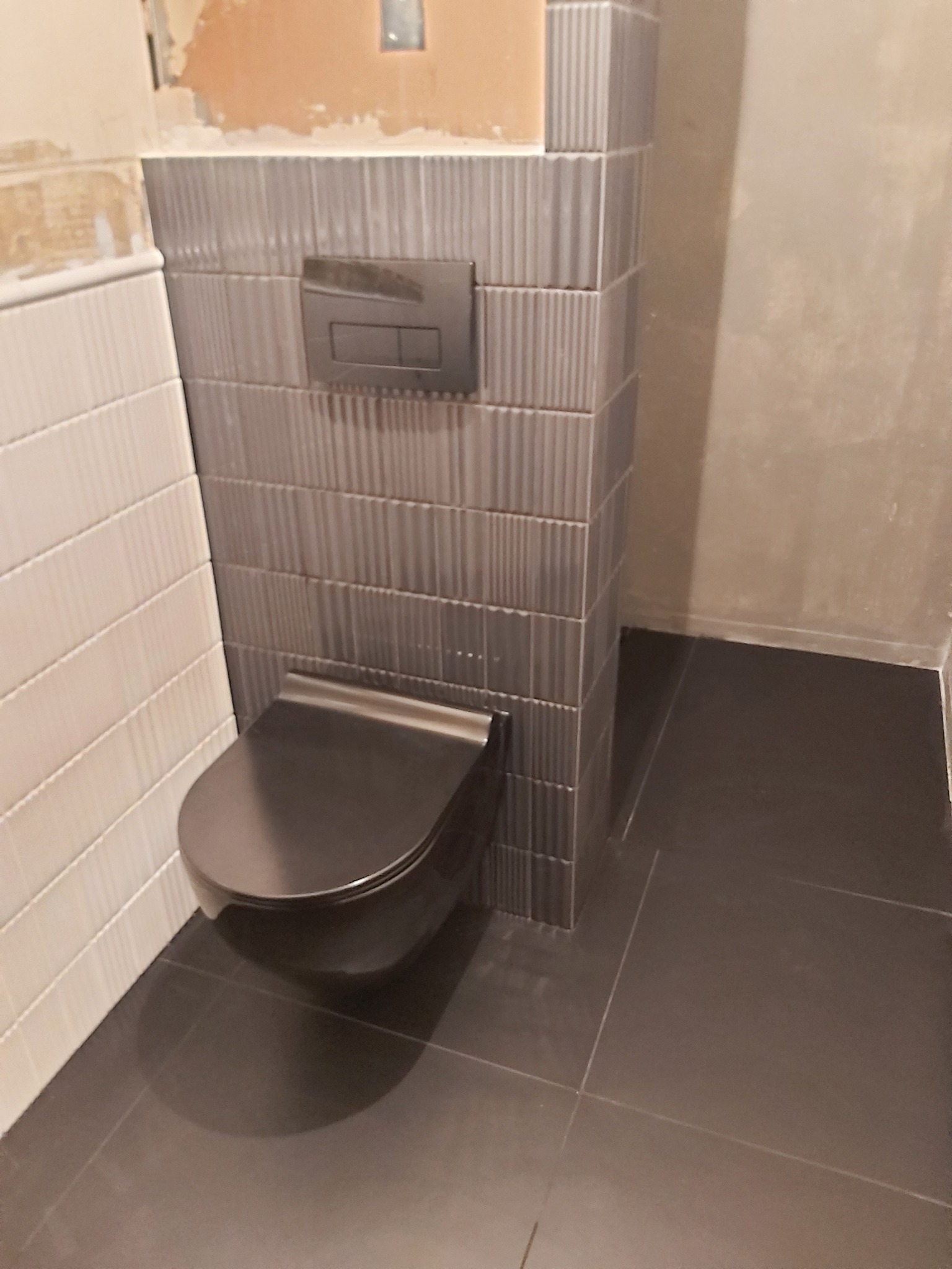 Nowoczesna toaleta podwieszana z szarą armaturą i płytkami w łazience. Widoczne białe i szare płytki na ścianach oraz ciemna podłoga. Częściowo wykończone wnętrze.