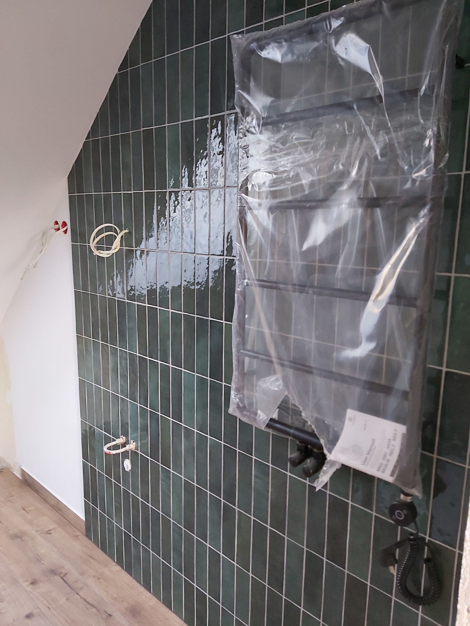 Łazienka w trakcie wykańczania: zielone, błyszczące płytki na ścianie, grzejnik drabinkowy w folii, widoczne instalacje elektryczne i hydrauliczne. Podłoga drewnopodobna.