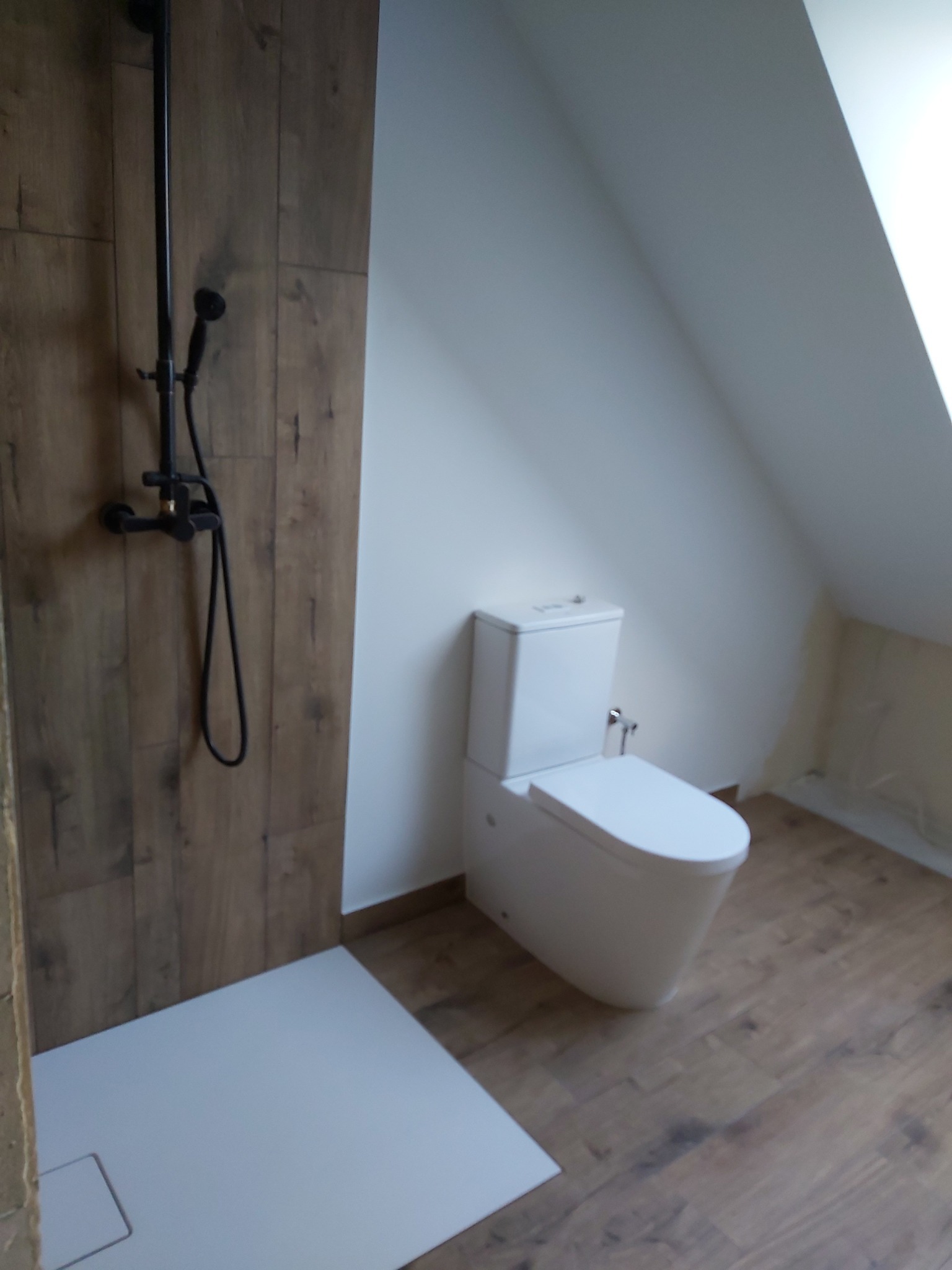 Nowoczesna łazienka na poddaszu z kabiną prysznicową typu walk-in, wykończona płytkami imitującymi drewno i białą ceramiką. Uwagę zwraca czarna armatura.