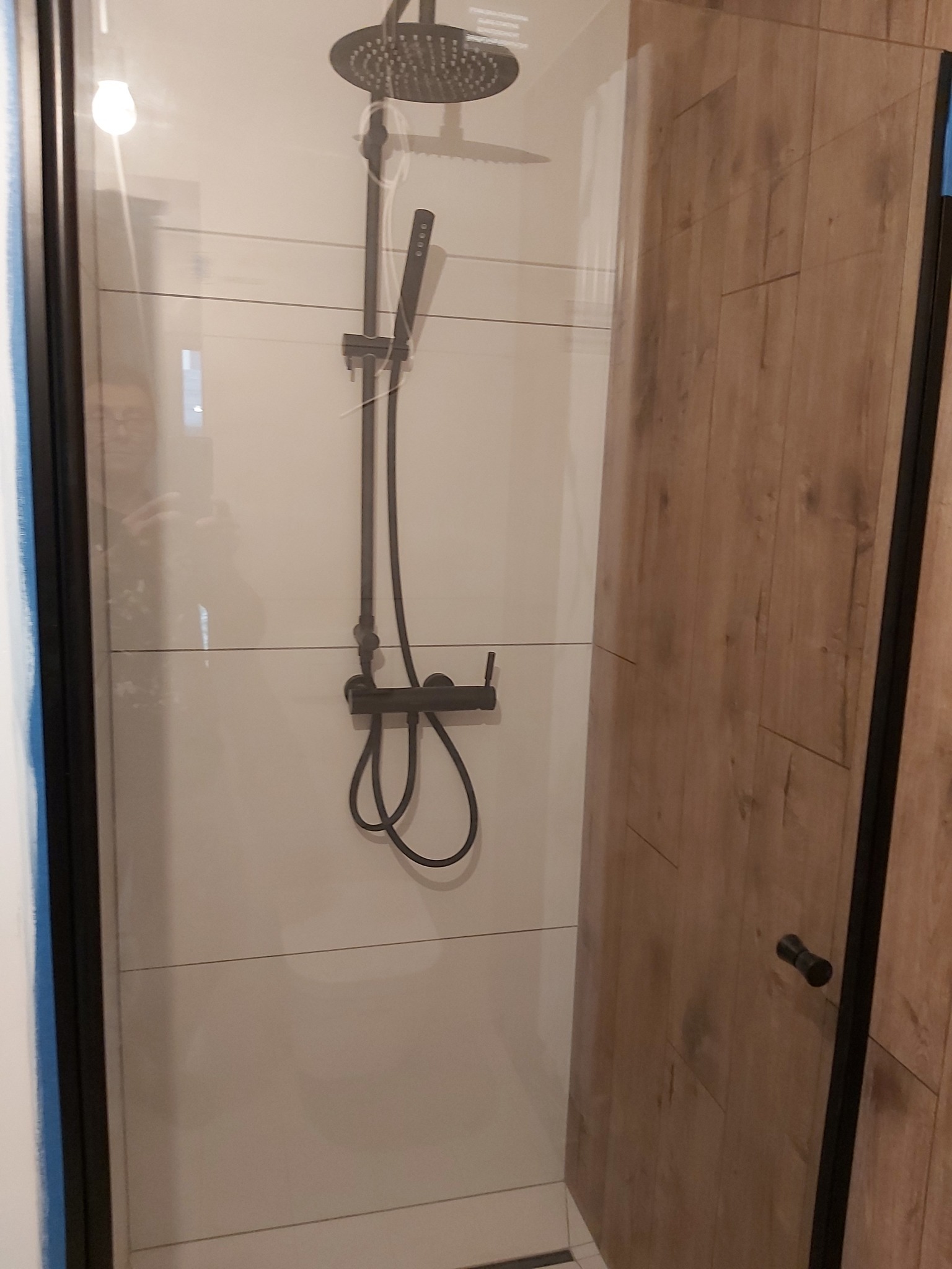 Nowoczesna kabina prysznicowa z czarną armaturą i drewnopodobnymi drzwiami, widok z boku, minimalistyczny design, jasne płytki na ścianach.