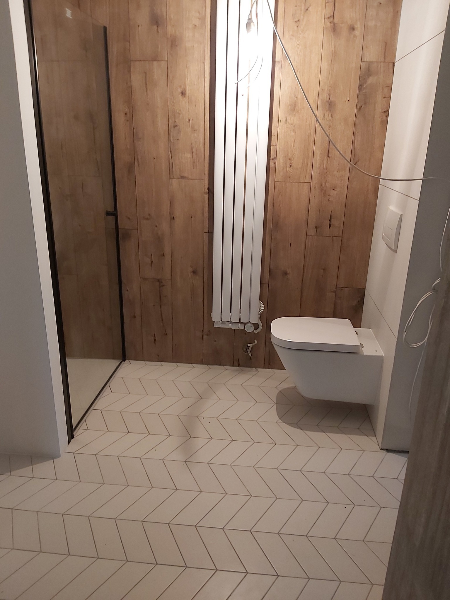 Nowoczesna łazienka z drewnopodobnymi panelami ściennymi, białą armaturą i podłogą w jodełkę. Kabina prysznicowa ze szklanymi drzwiami i grzejnik drabinkowy.