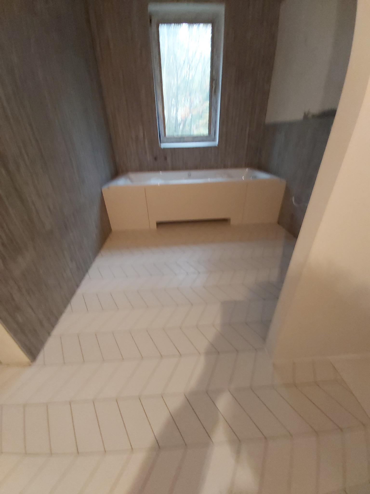 Jasna łazienka na poddaszu z oknem. Białe płytki ułożone w jodełkę na podłodze, zabudowana wanna. Ściany wykończone w odcieniach szarości. Ujęcie z perspektywy podłogi.