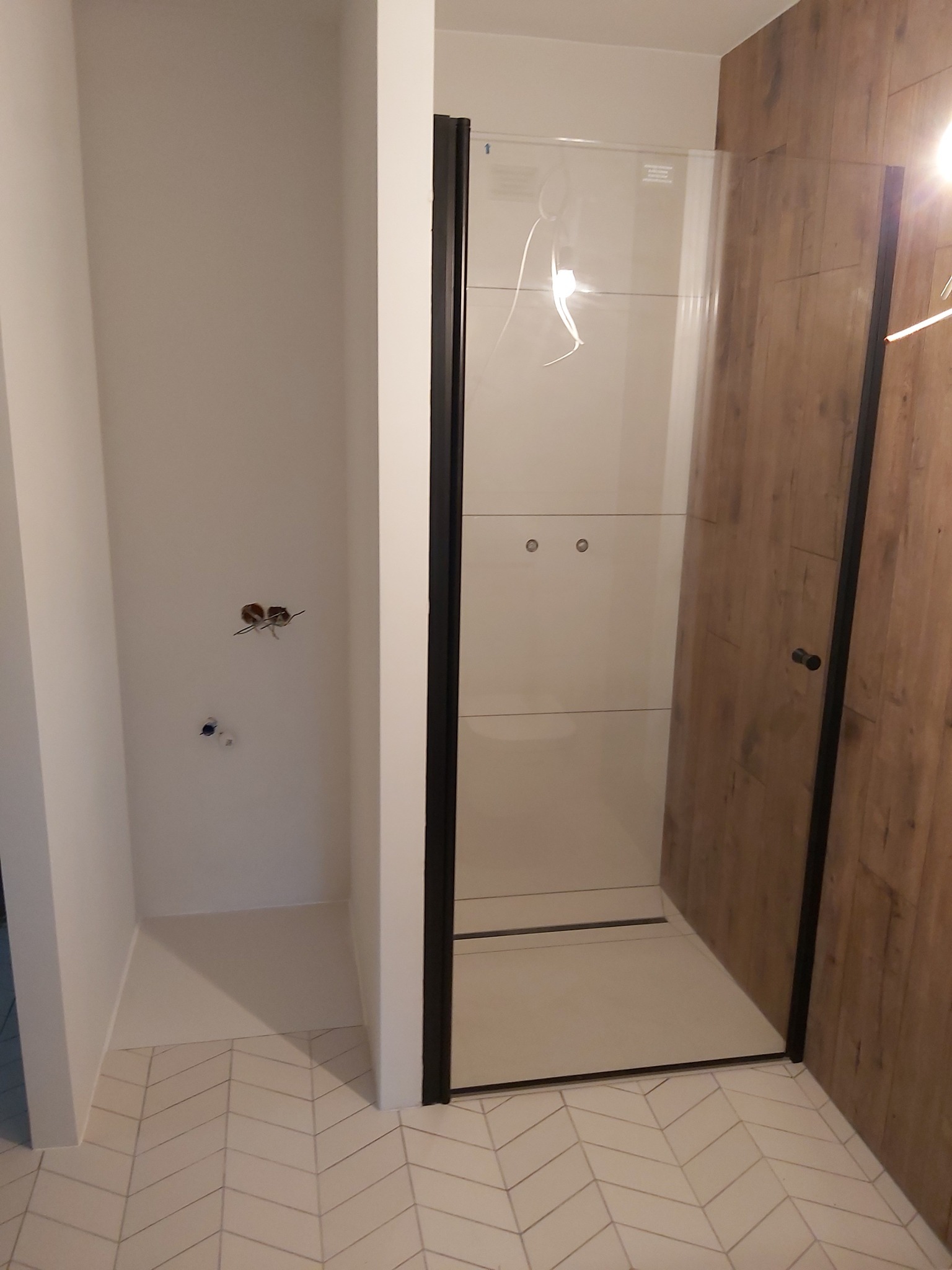 Nowoczesna kabina prysznicowa z czarnymi profilami, jasne płytki podłogowe ułożone w jodełkę, drewnopodobne panele na ścianie. Widoczne przygotowane punkty instalacji.