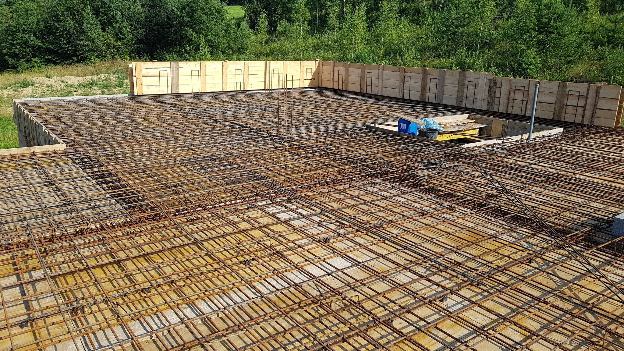 Zbrojenie stropu budynku jednorodzinnego z widocznymi szalunkami i wystającymi prętami. Prace budowlane w toku, widok z góry na siatkę zbrojeniową.