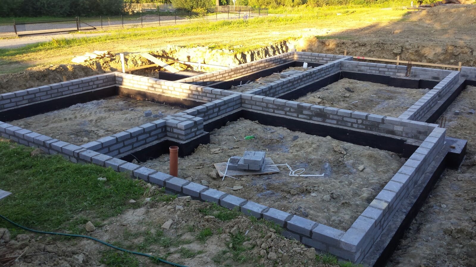 Wykopane fundamenty z izolacją i częściowo wymurowanymi ścianami z szarych bloczków. Widoczne wystające rury i pozostałości materiałów budowlanych.