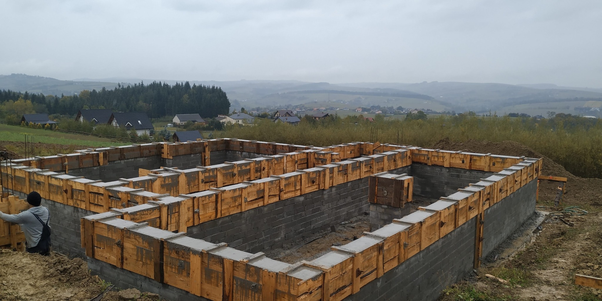 Fundamenty domu jednorodzinnego z szalunkiem drewnianym na tle mglistego krajobrazu. Widoczny pracownik w trakcie wykonywania prac.