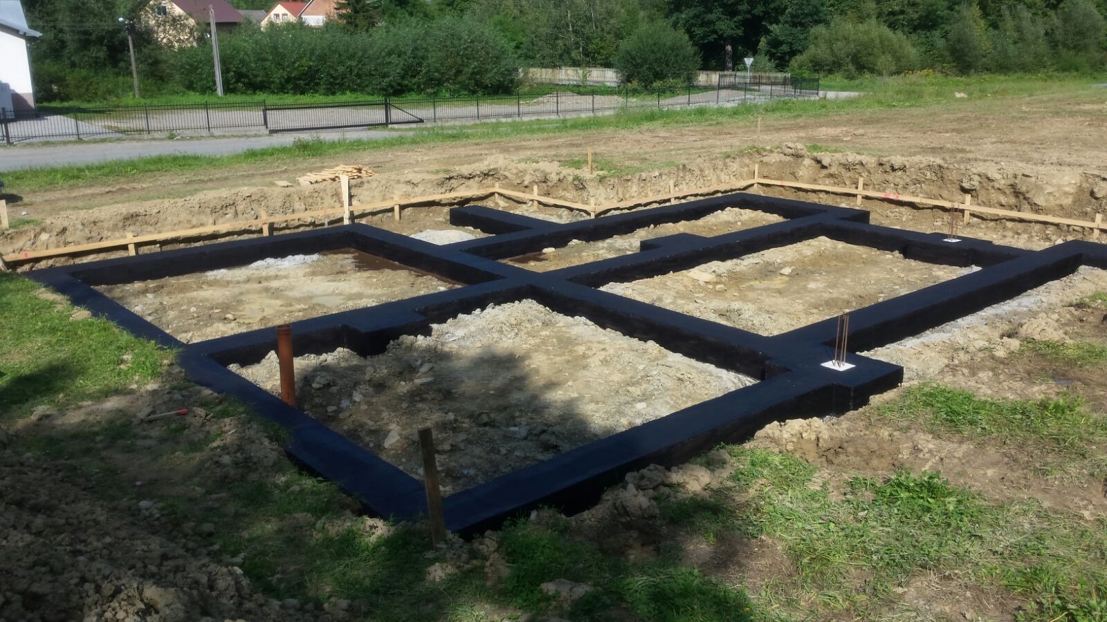 Zalane fundamenty domu, zabezpieczone czarną izolacją, widoczne drewniane szalunki i wystające pręty zbrojeniowe. Tło: zielona trawa, drzewa, domy.