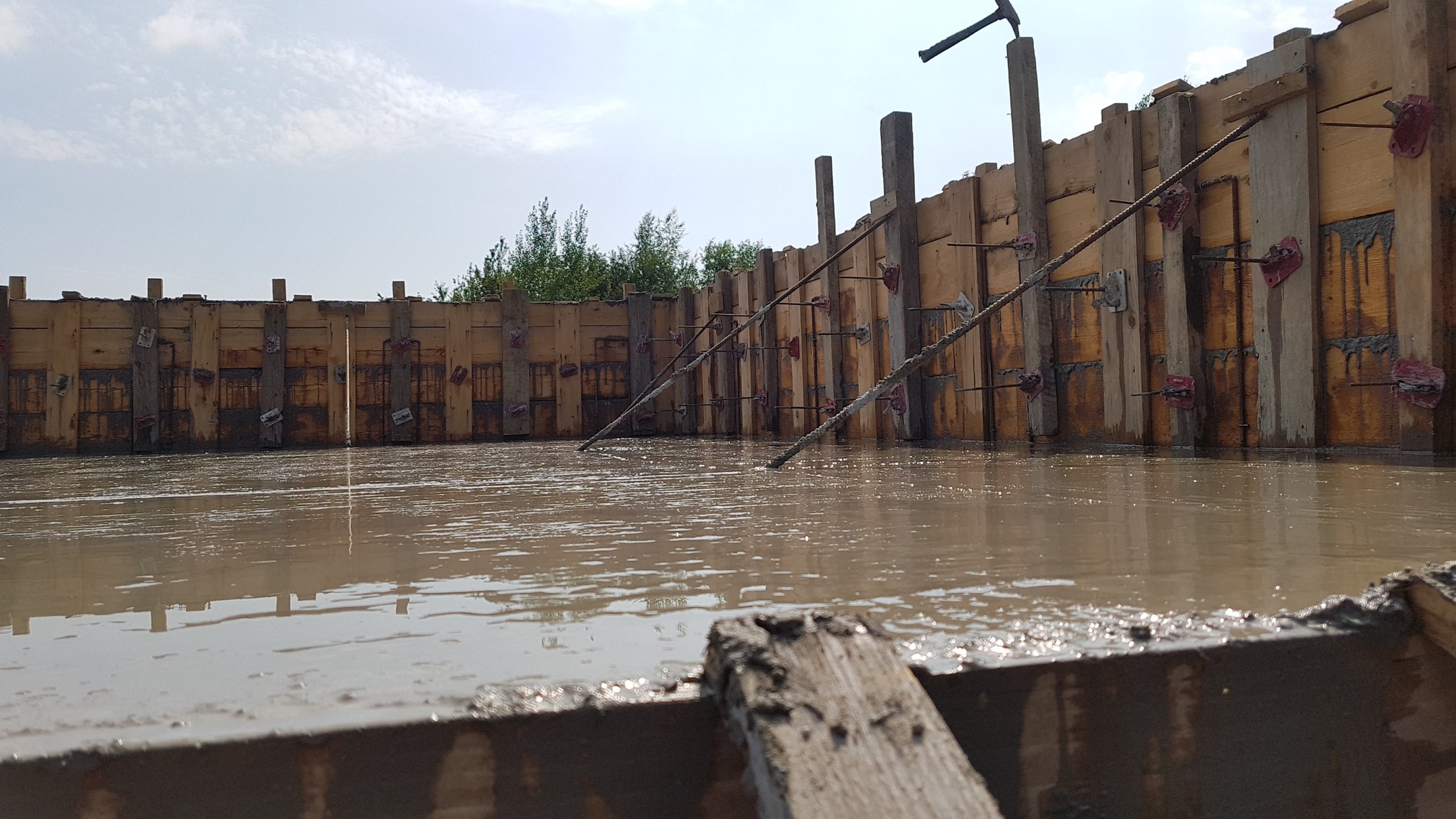 Zalany fundament z drewnianym szalunkiem wzmocnionym stalowymi prętami. Widoczne ślady betonu na deskach. W tle zieleń drzew i błękitne niebo.