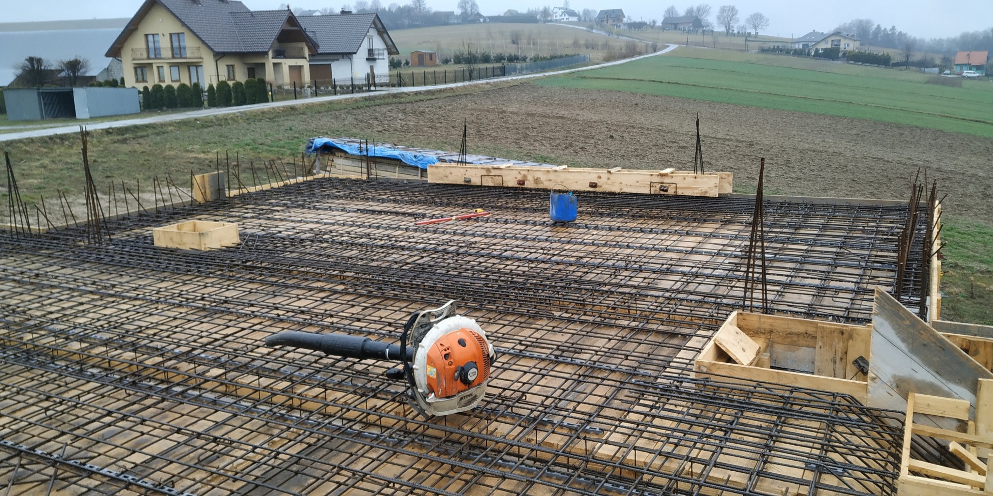 Zbrojenie fundamentów pod budynek w Posadowej Mogilskiej. Widoczna siatka z prętów, szalunki i dmuchawa do czyszczenia. Prace budowlane w toku.