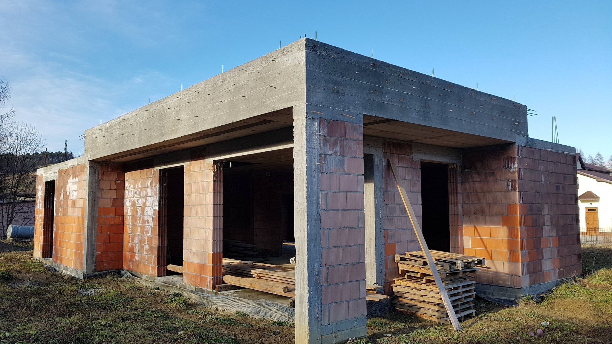 Surowy stan budynku w trakcie budowy: ściany z czerwonej cegły, betonowy strop, drewniane palety. Widoczne zbrojenia i otwory okienne. Błękitne niebo w tle.