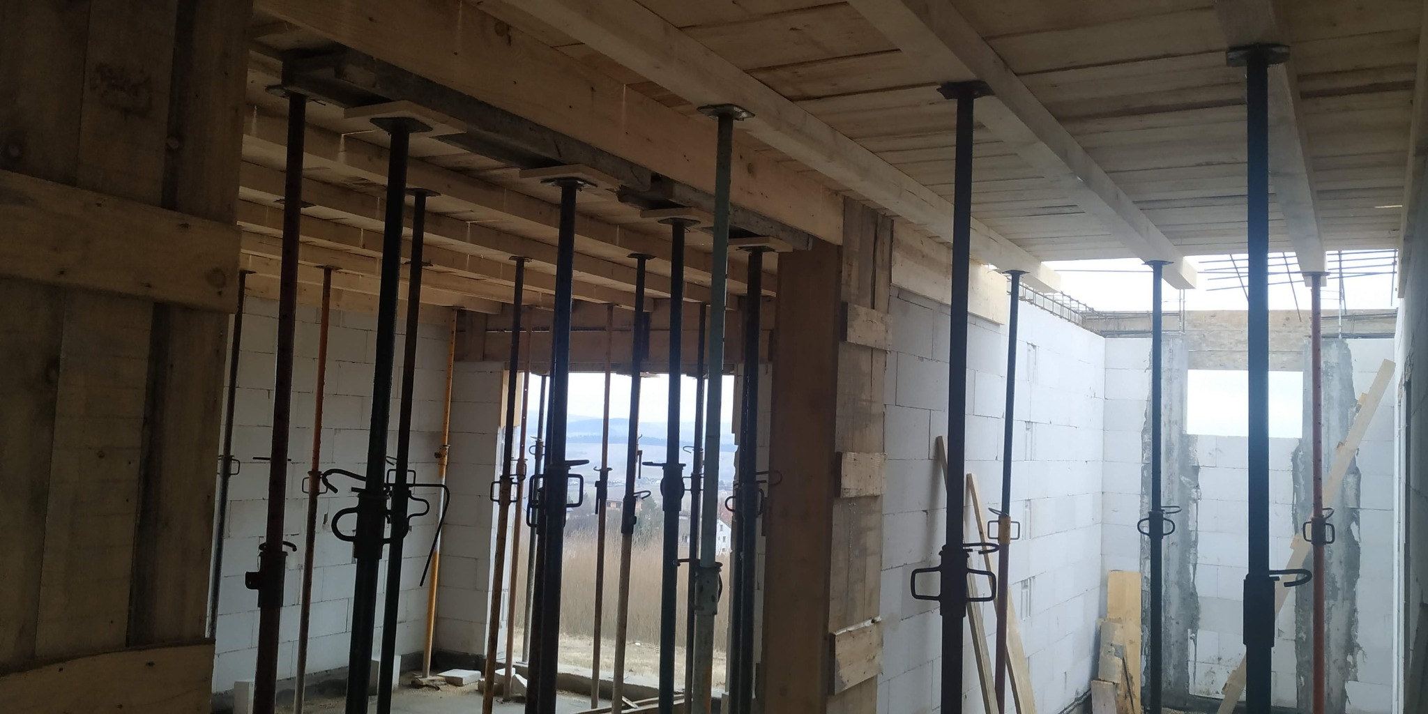 Wnętrze w trakcie budowy: widoczne podpory stropowe, ściany z betonu komórkowego i drewniane elementy konstrukcyjne. W tle widok na krajobraz.