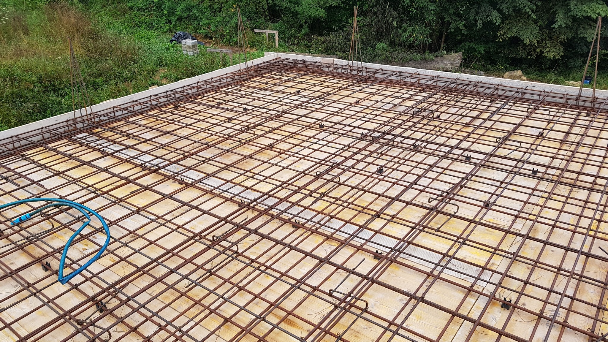 Zbrojenie płyty fundamentowej z widoczną siatką stalową, drewnianym szalunkiem i fragmentem niebieskiego węża. Widok z góry na placu budowy.