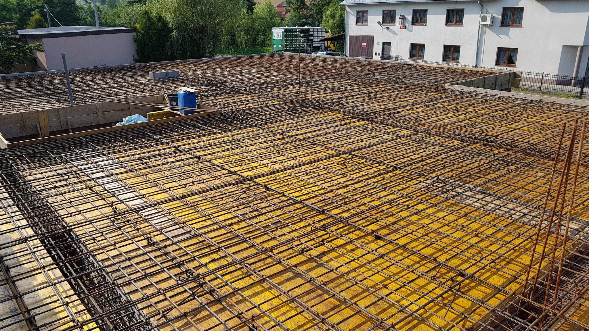 Zbrojenie stropu na żółtym szalunku, widok z góry. Stalowe pręty tworzą regularną siatkę. W tle budynek z białej cegły i zieleń drzew.