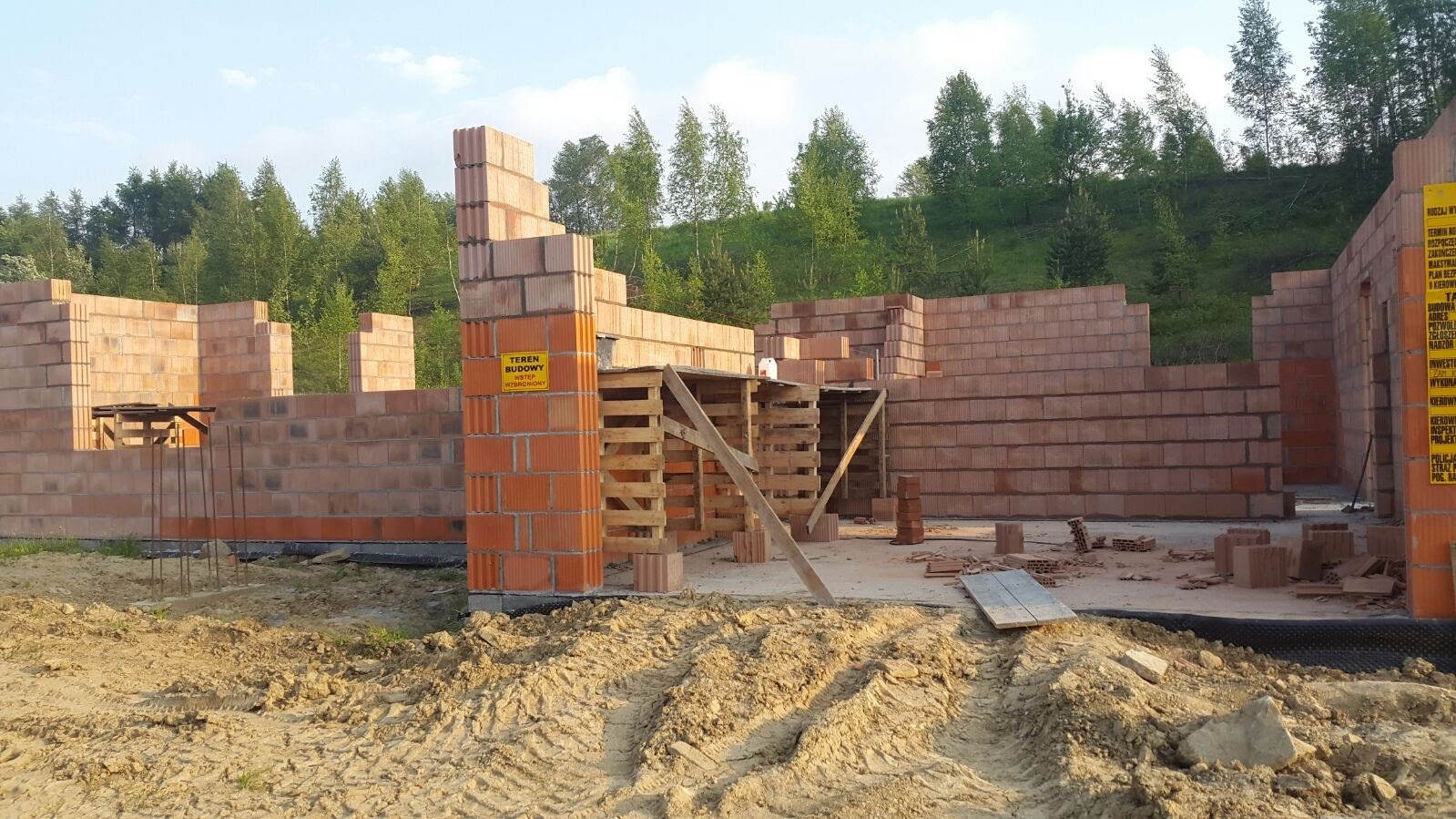 Budowa domu w trakcie wznoszenia ścian z czerwonej cegły. Widoczny teren budowy z tablicą informacyjną i fragment lasu w tle. Ujęcie z poziomu gruntu.