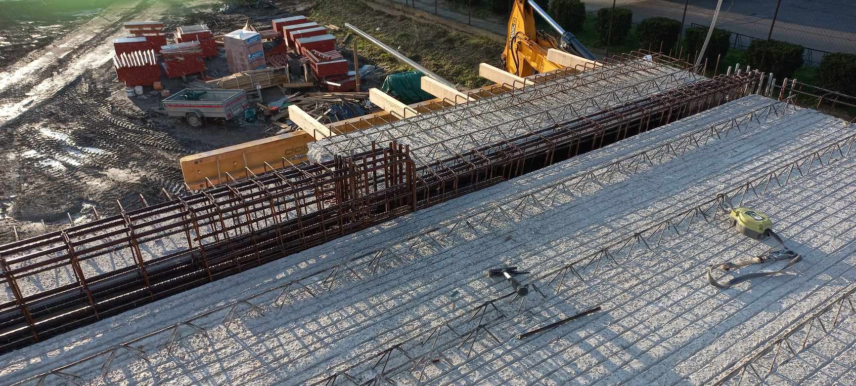 Zbrojenie fundamentów pod budowę z widocznymi elementami szalunków i sprzętem budowlanym w tle. Ujęcie z góry ukazuje detale konstrukcji.